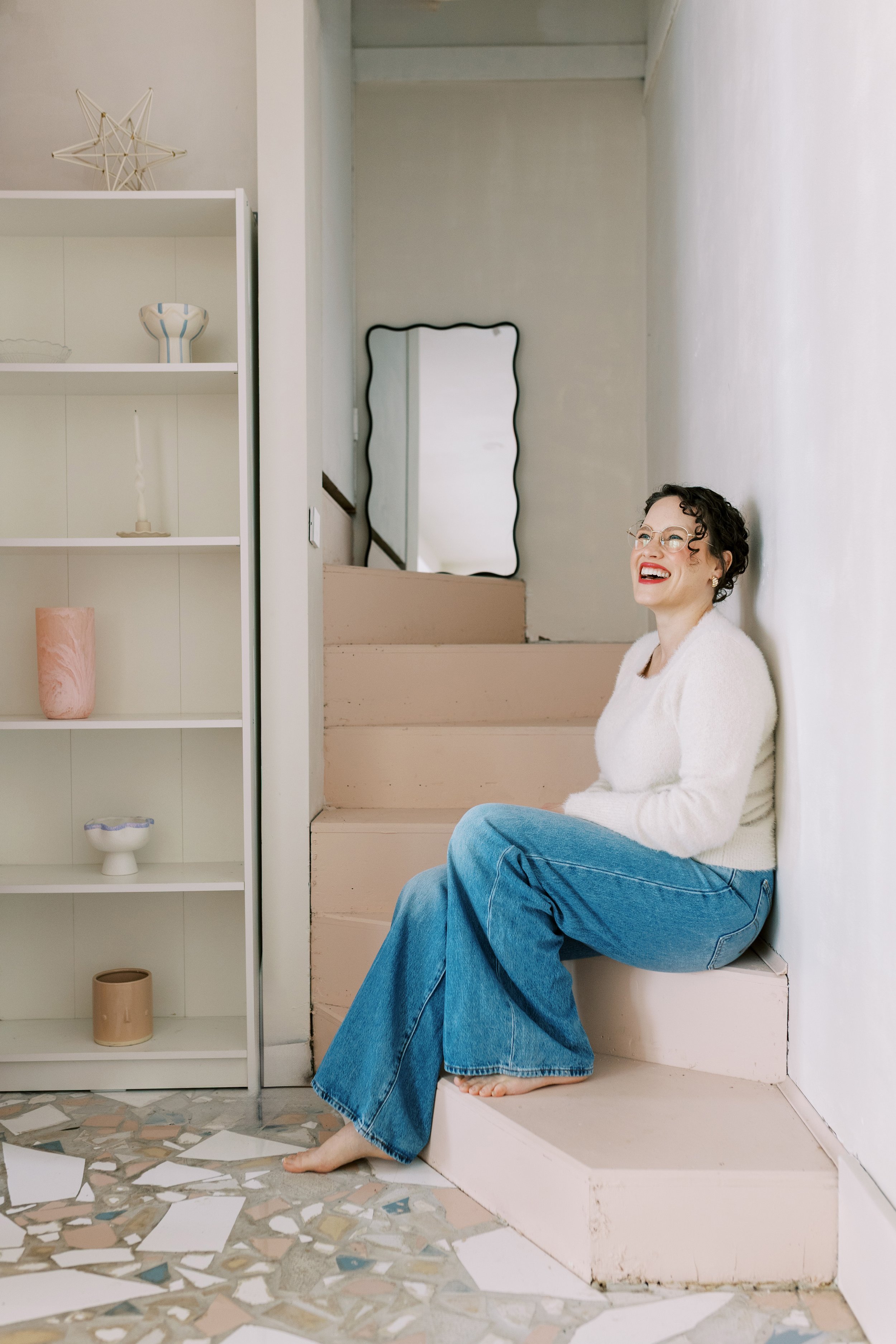A woman with short curly hair, glasses, red lipstick, and a white sweater sitting barefoot on a pink staircase, smiling and leaning against a white wall.