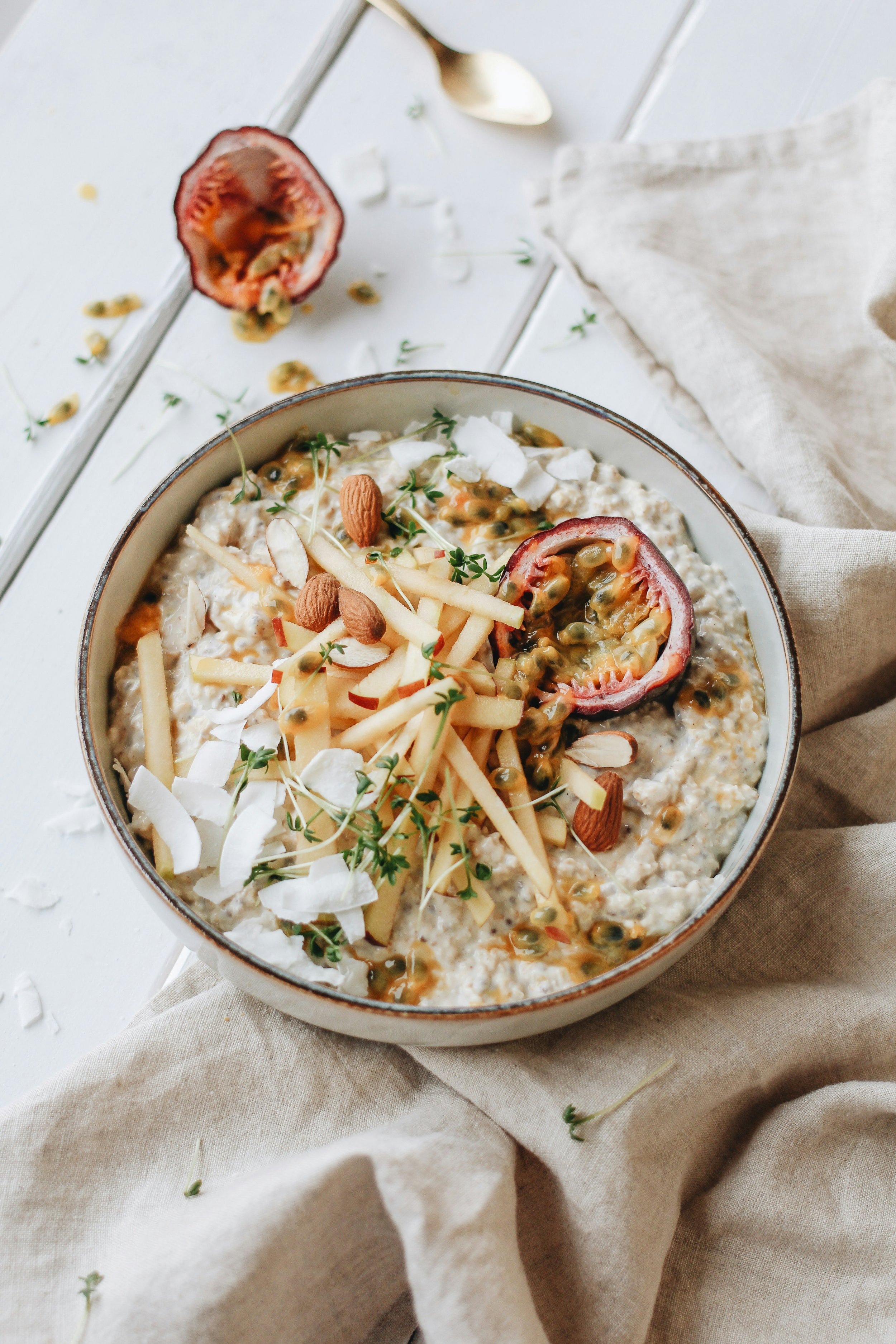 A bowl of oatmeal topped with sliced almonds, shredded cheese, almonds, microgreens, passion fruit halves, and passion fruit seeds on a white textured surface with a spoon and a beige napkin.