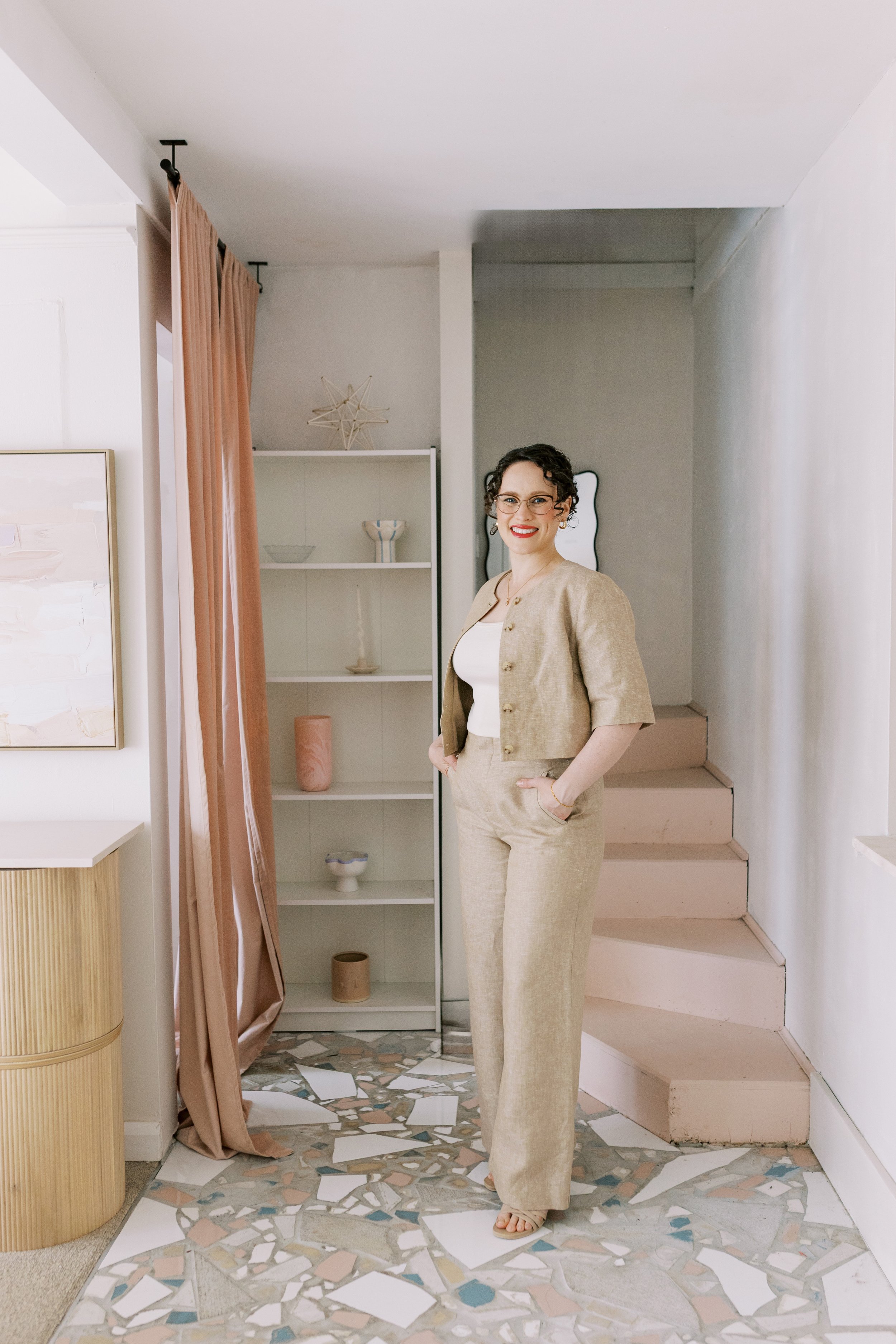 A woman with curly hair and glasses wearing a beige suit and white top, standing inside a room with modern decor, near a set of pink stairs and a white bookshelf with decorative objects.