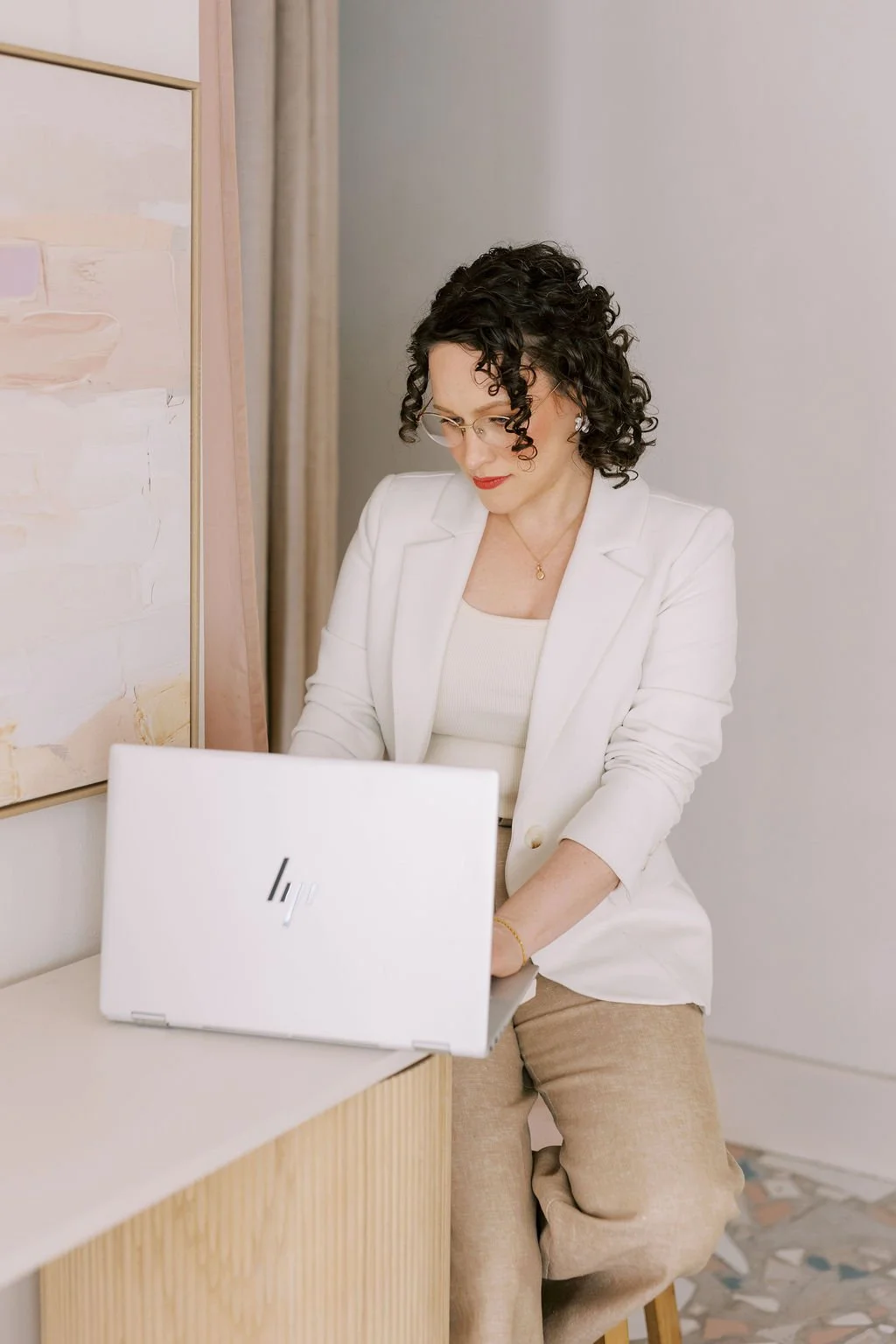 A woman with curly dark hair wearing glasses, a white blazer, and beige pants working on a white HP laptop in a modern, minimalistic room with a textured painting and patterned flooring.