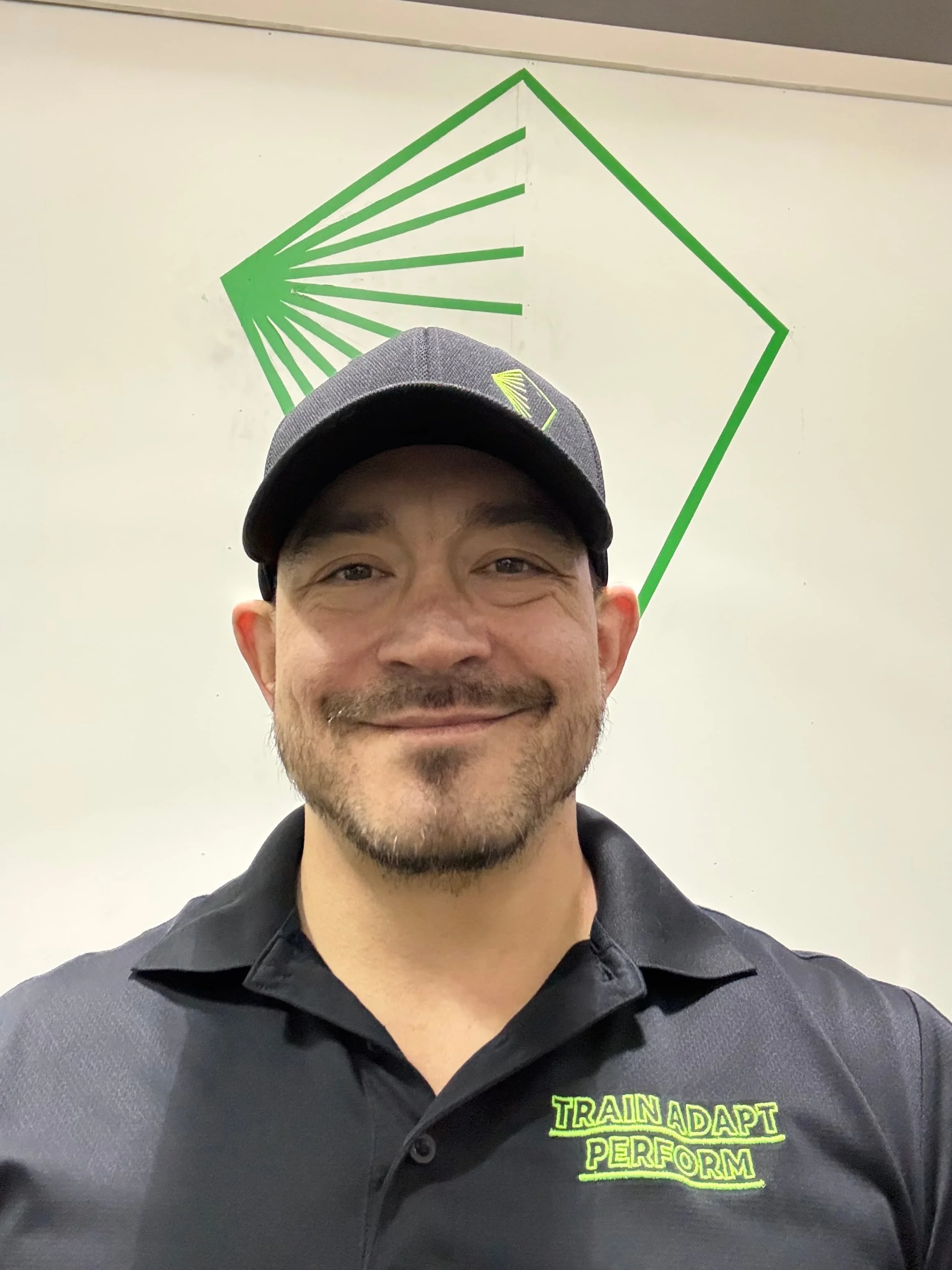 A smiling man wearing a black polo shirt with neon green text that says 'Train Adapt Perform' and a grey baseball cap with a yellow logo. Behind him is a white wall with a green geometric design.
