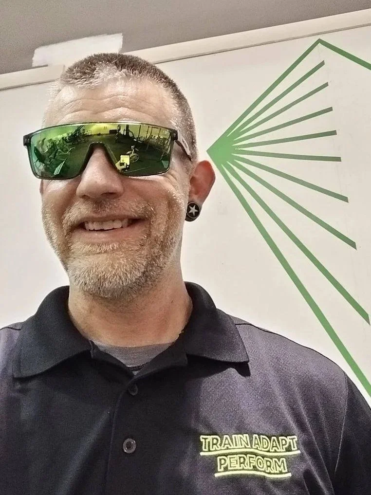 Man with short hair, sunglasses, and facial hair smiling in front of a white wall with green graphic design. Wearing a black polo shirt with yellow text that reads 'Train, Adapt, Perform.'