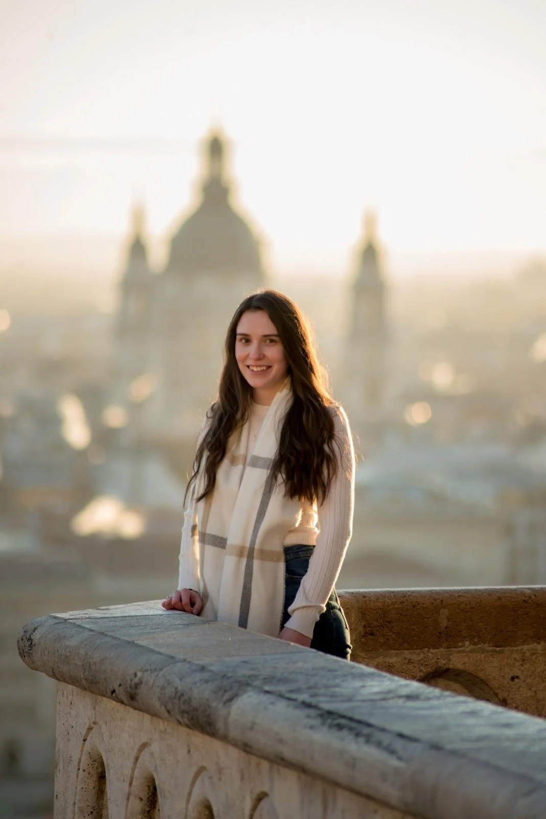 A young woman with long dark hair smiling, standing outdoors on a bridge with a stone railing, in front of a blurred cityscape with historic architecture, during golden hour.