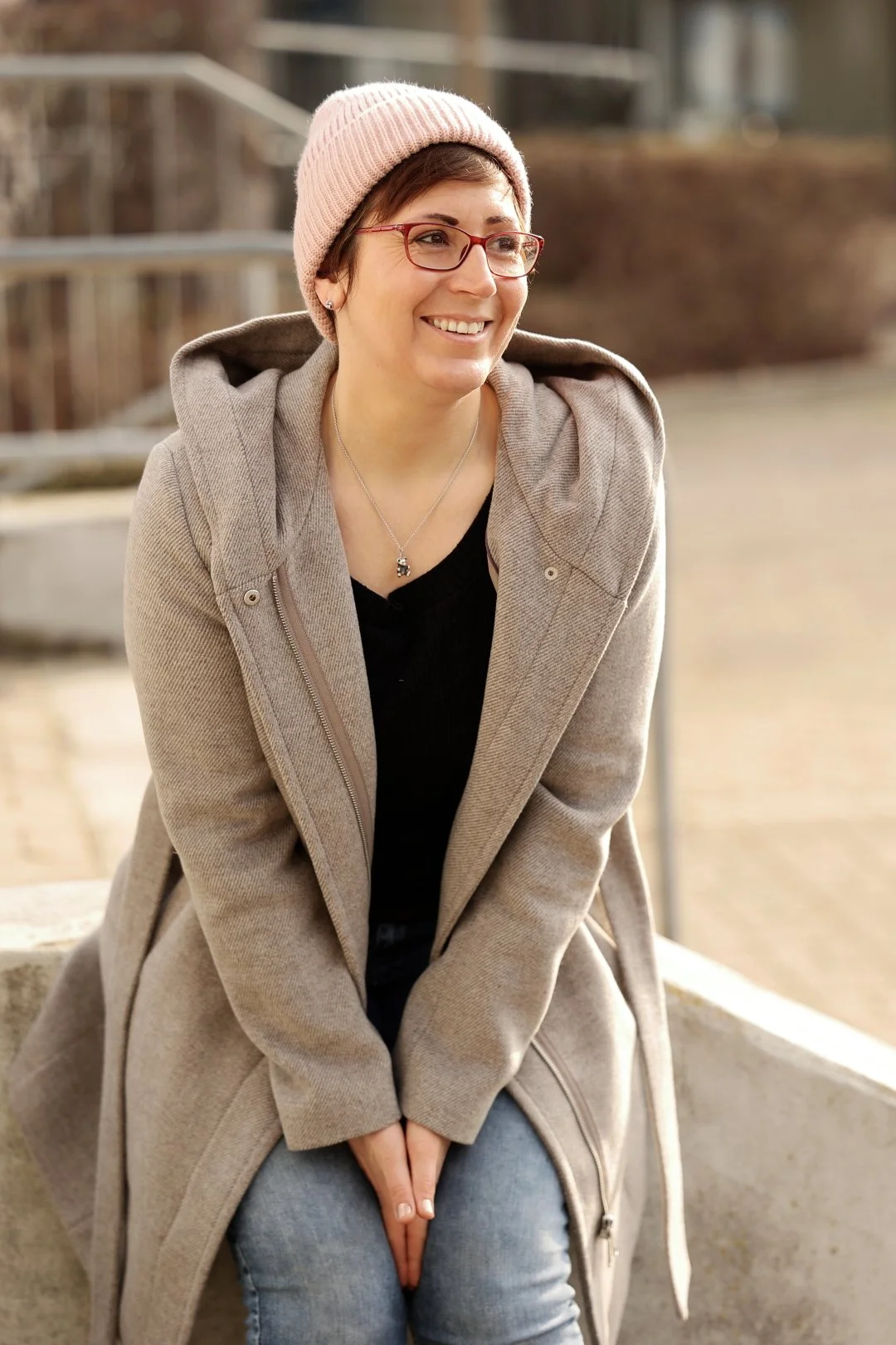 Eine lächelnde Frau mit Brille trägt einen rosa Wollmütze, einen beigen Mantel und sitzt auf einer Bank im Freien.