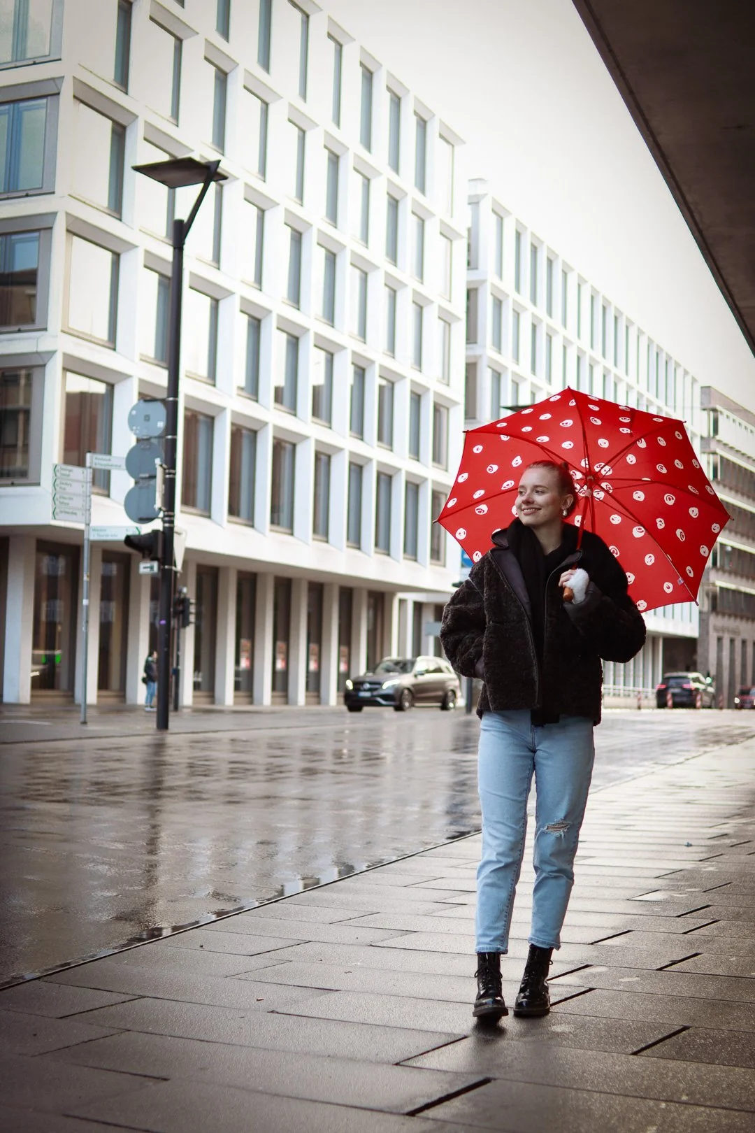Eine junge Frau läuft an einem regnerischen Tag auf einem Stadtplatz, hält einen roten Regenschirm mit weißen Mustern und trägt eine schwarze Jacke, Jeans und schwarze stiefel.