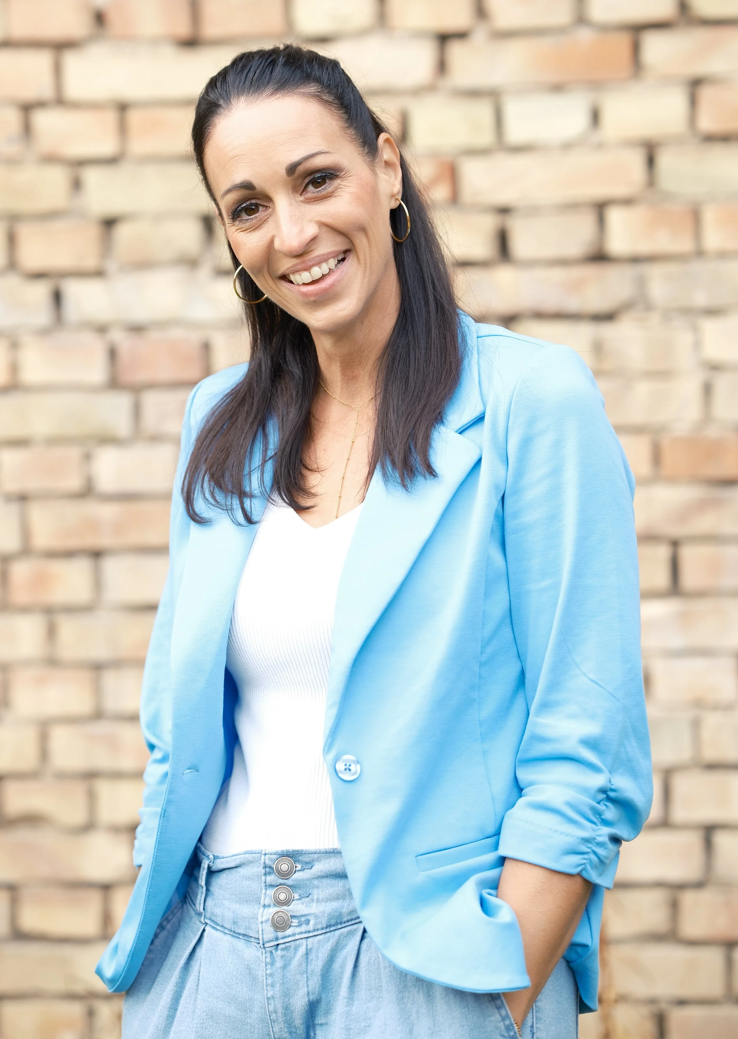 Eine lachende Frau mit dunklem, langem Haar, trägt eine hellblaue Jacke, weiße Top und hellblaue Jeans, steht vor einer Ziegelmauer.