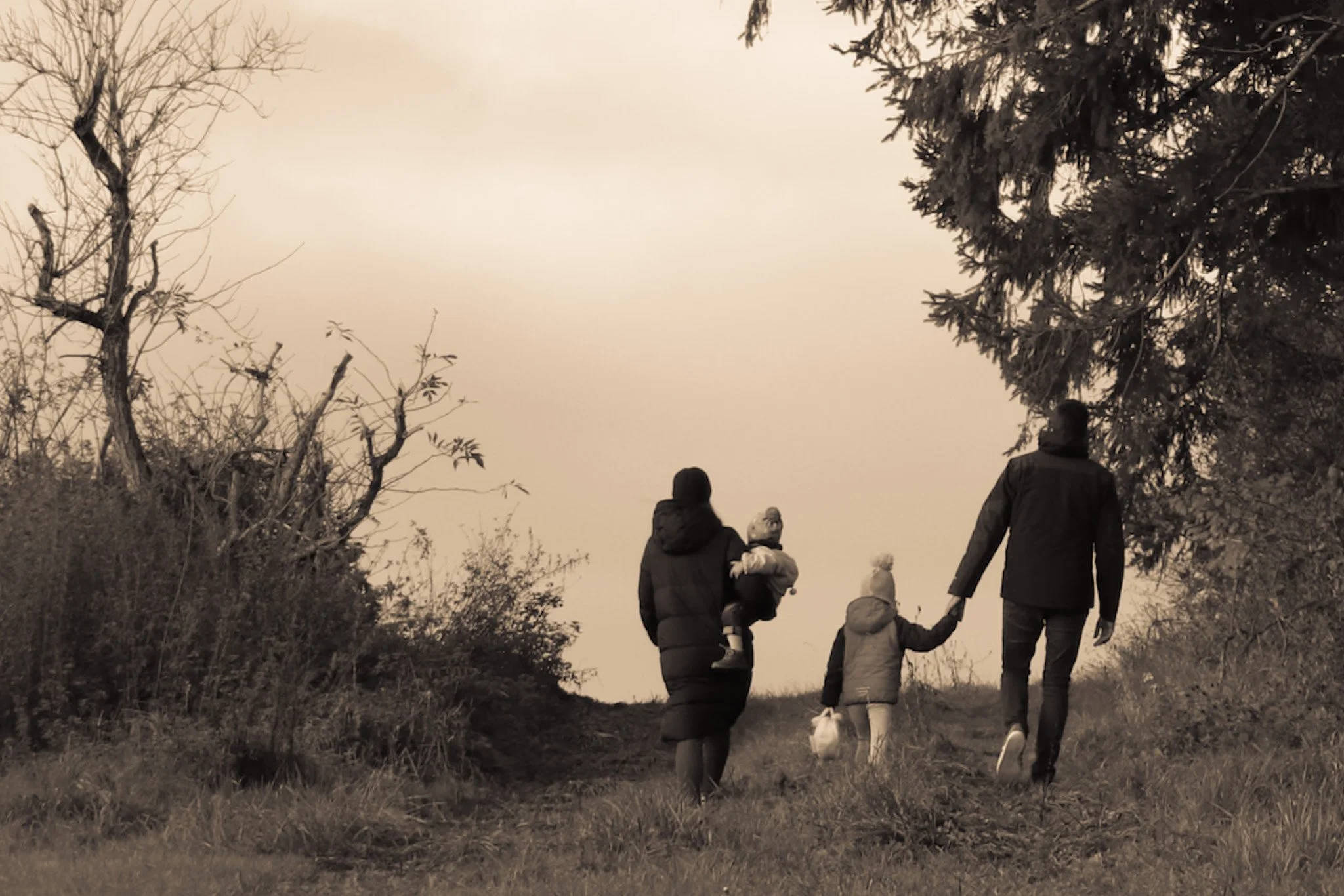 Familie mit zwei Kindern geht auf einem Weg im Freien, umgeben von Bäumen, bei Sonnenuntergang.