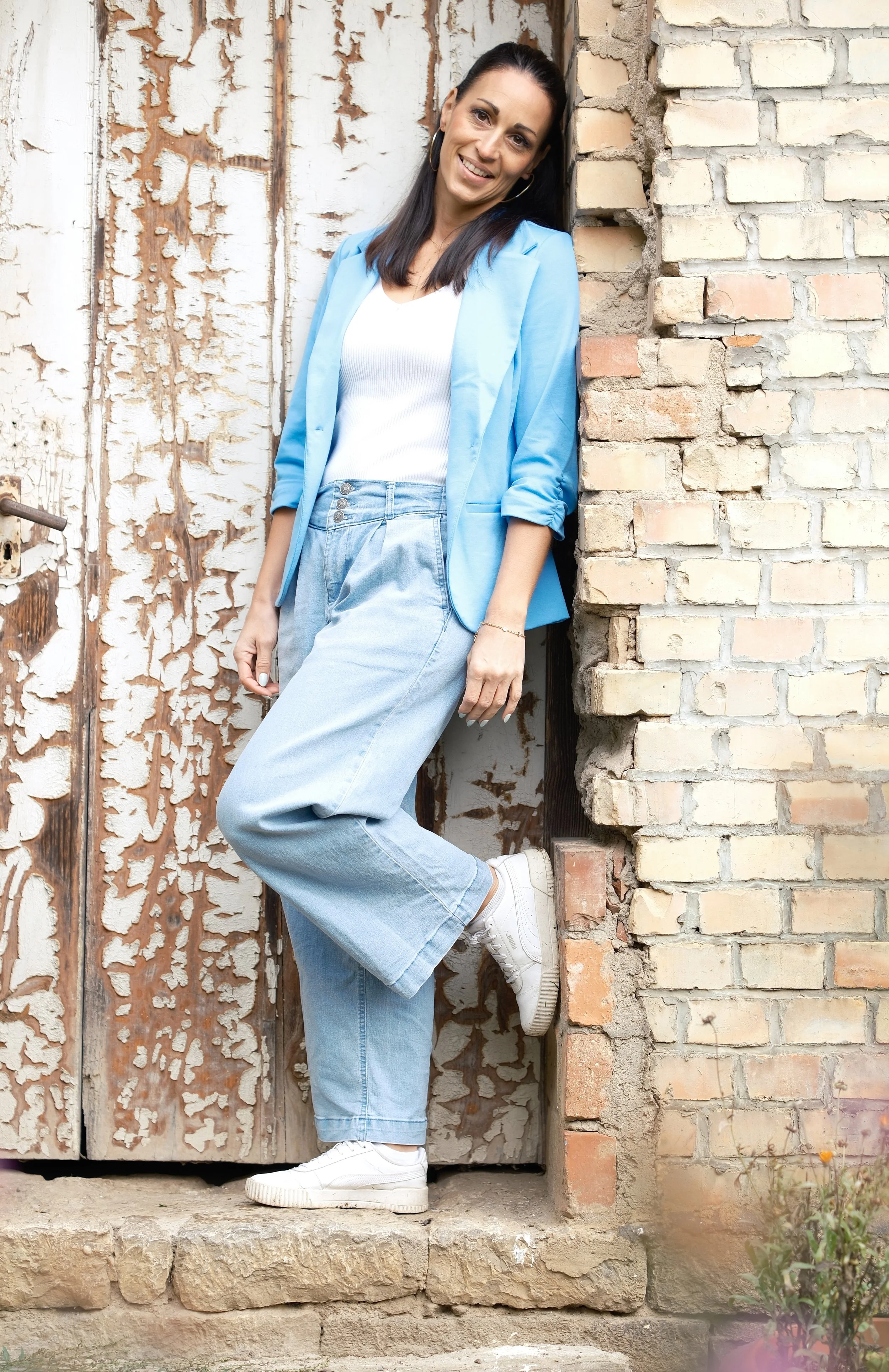 Eine Frau steht neben einer alten, bemalten Tür und einer Ziegelmauer, trägt eine blaue Blazer, eine weiße Top, weite helle Jeans und weiße Turnschuhe, lächelt.