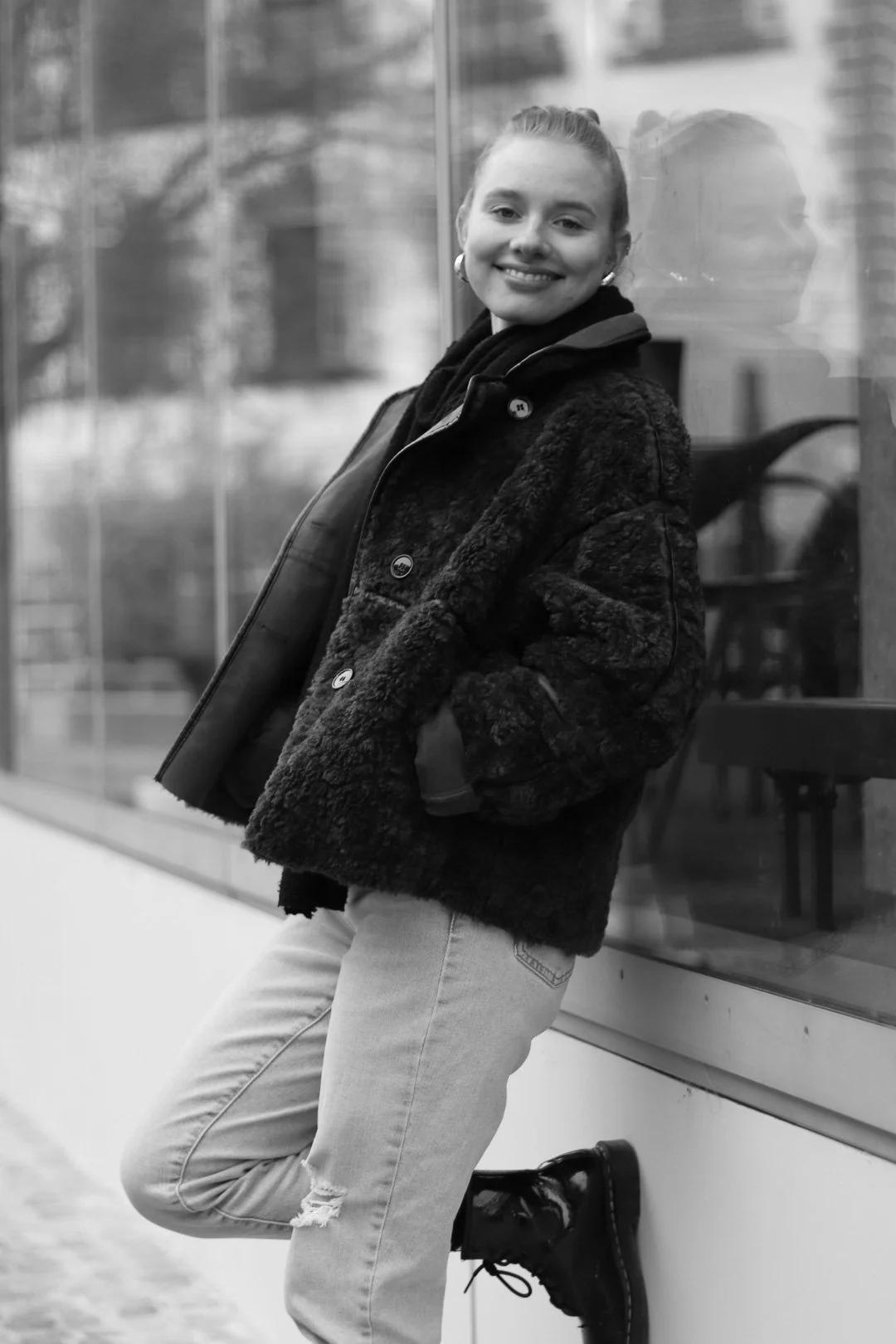 Ein lächelndes junges Mädchen in einem dunklen, pelzartigen Mantel und hellen Jeans, das an einer Glaswand steht und auf die Kamera schaut.