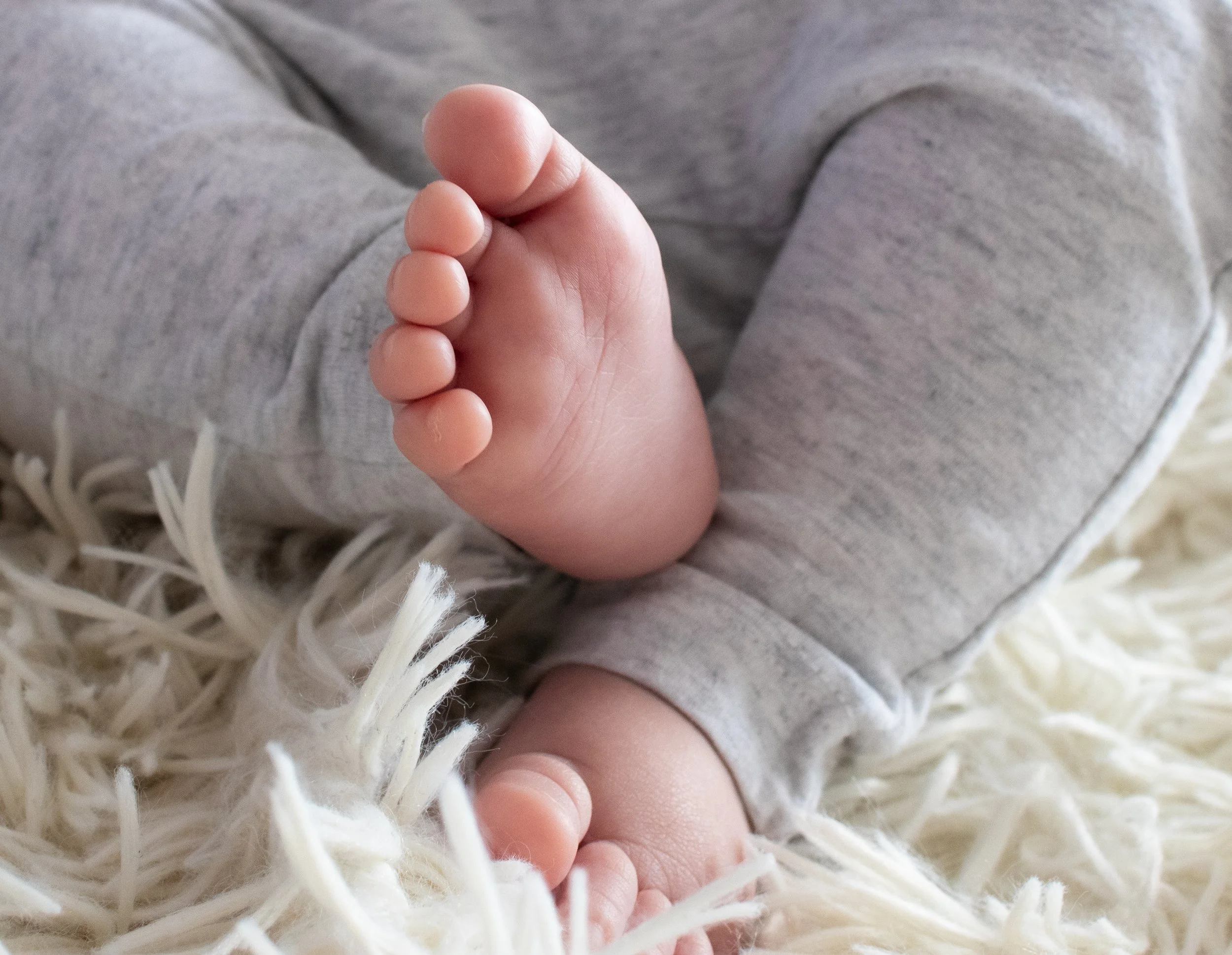 Nahaufnahme eines Babyfußes, das auf einem weichen, cremefarbenen Teppich liegt, während das Baby in grauem Kleidung steckt.