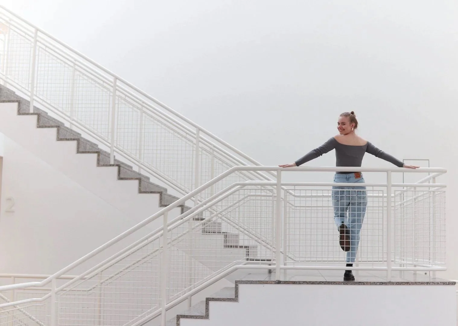 Eine lachende junge Frau steht auf einer weißen Treppe in einem modernen, hellen Gebäude. Sie trägt ein graues Oberteil, Jeans und schwarze Schuhe.