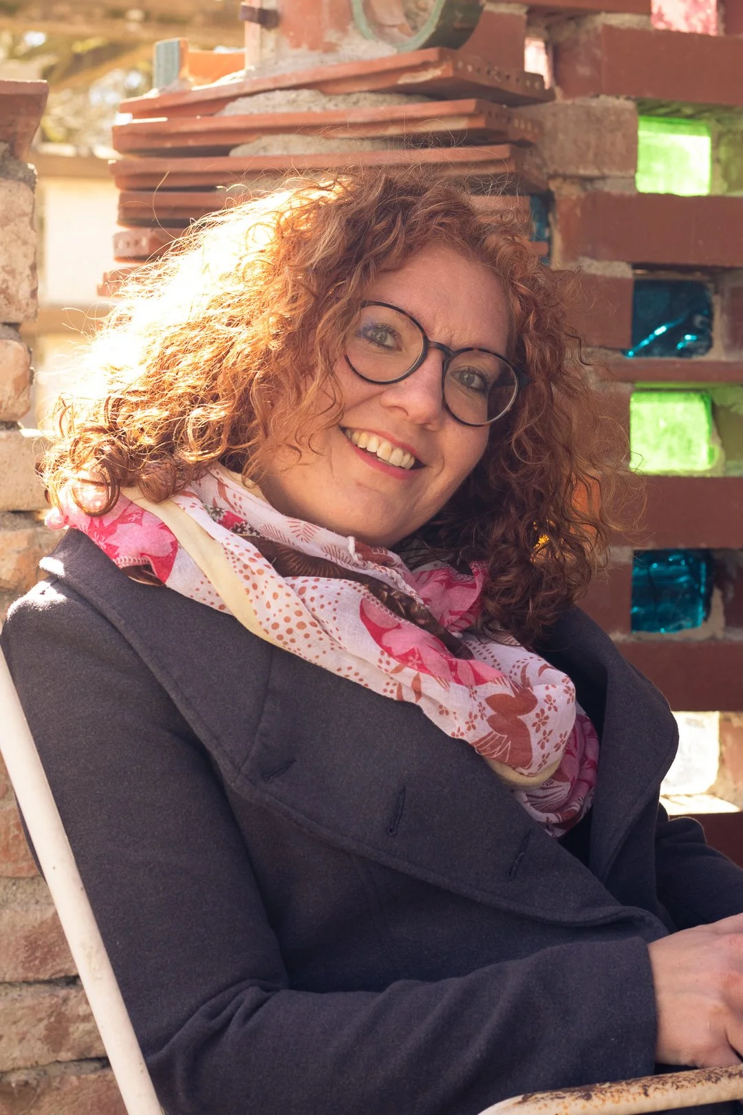 Eine lachende Frau mit lockigem, rotem Haar, trägt Brille und einen bunten Schal, sitzt vor einem Backsteinmauer. Es ist sonnig draußen.