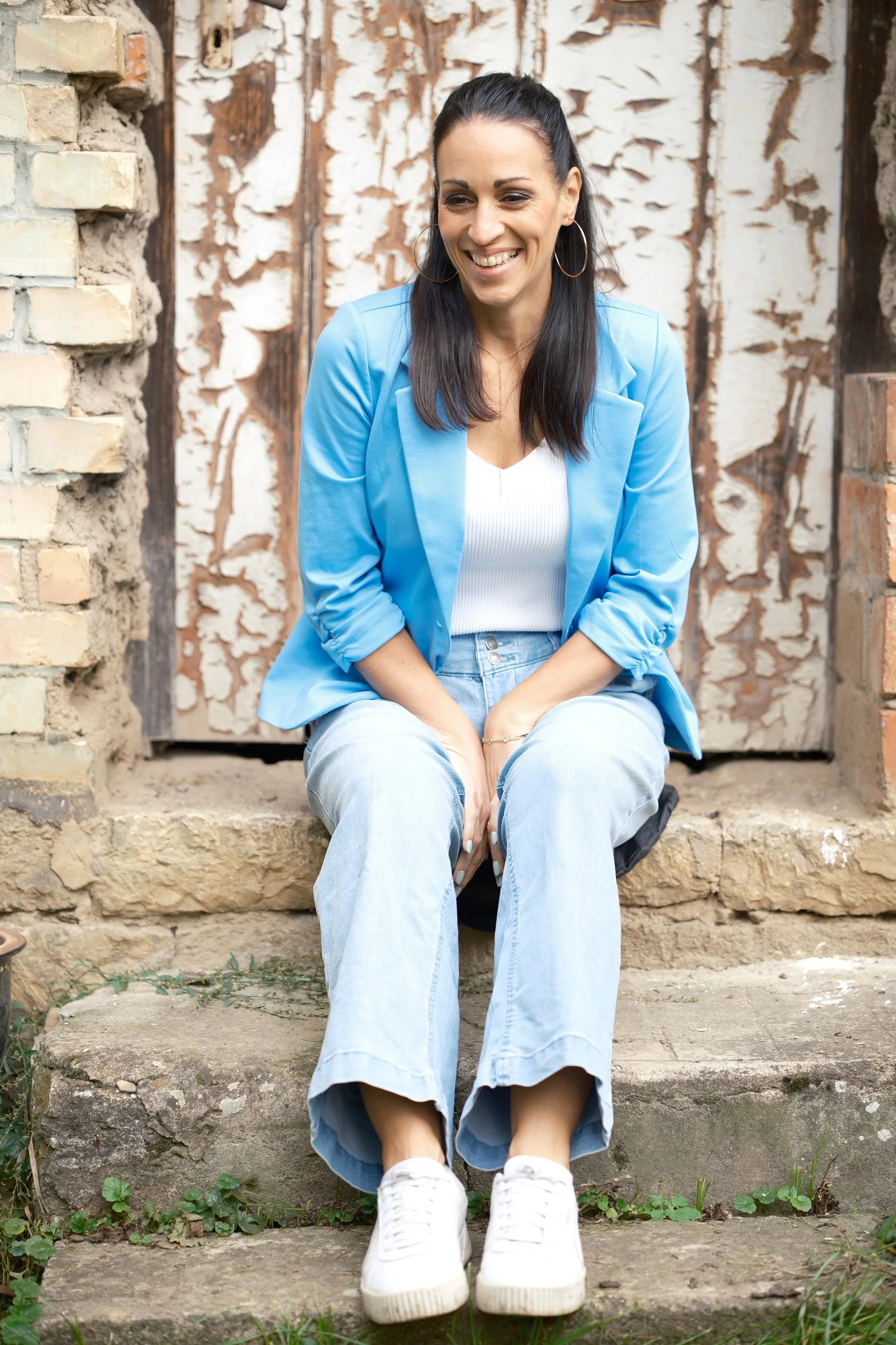 Eine lächelnde Frau sitzt auf einer Steintreppe vor einer alten, verzierten Holz- und Ziegelwand.