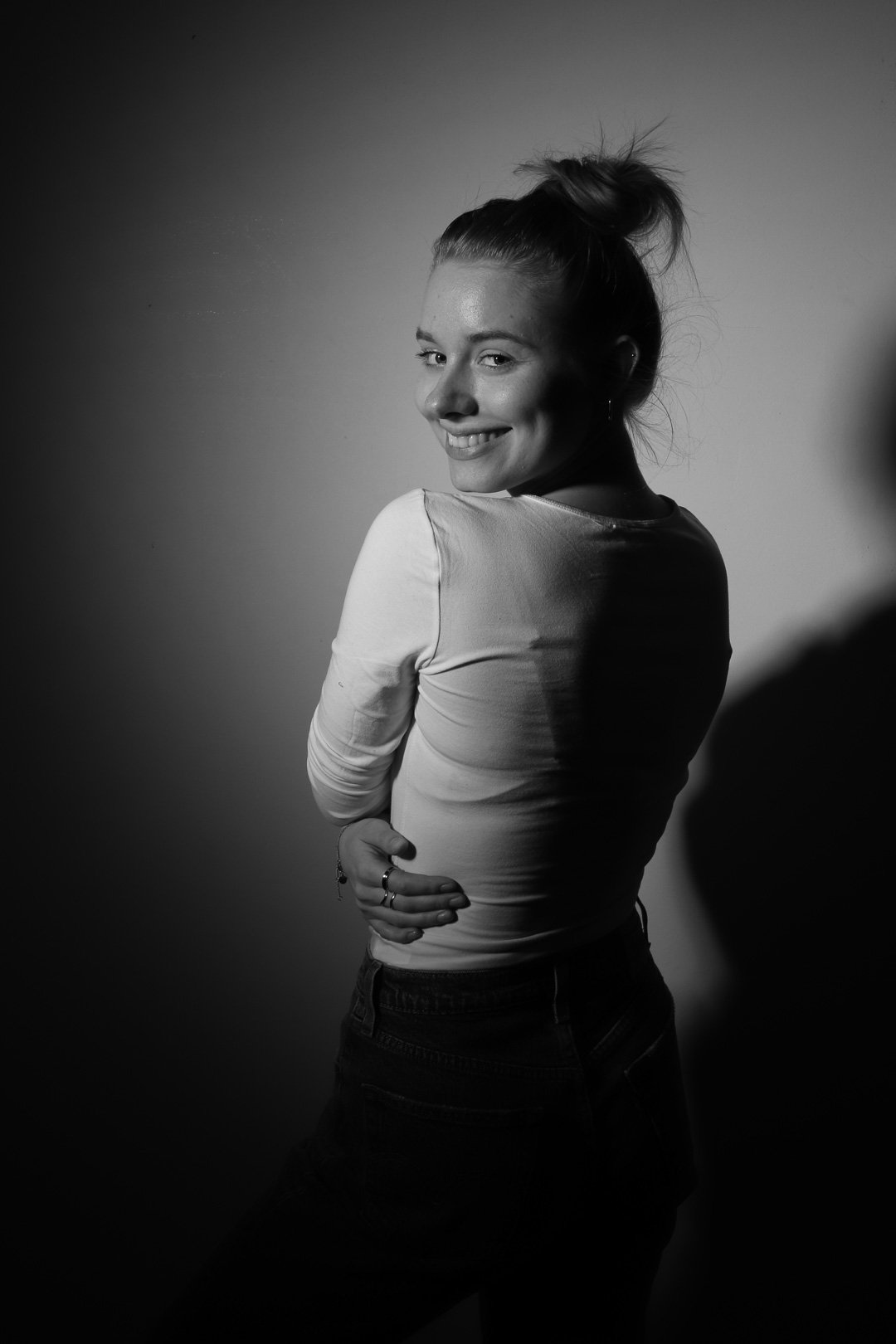 Schwarz-weiß-Fotografie eines lächelnden jungen Mädchens, das sich vor einer hellen Wand befindet, mit einem lockeren Pferdeschwanz und ördnungsspielen Schmuck an der Hand.