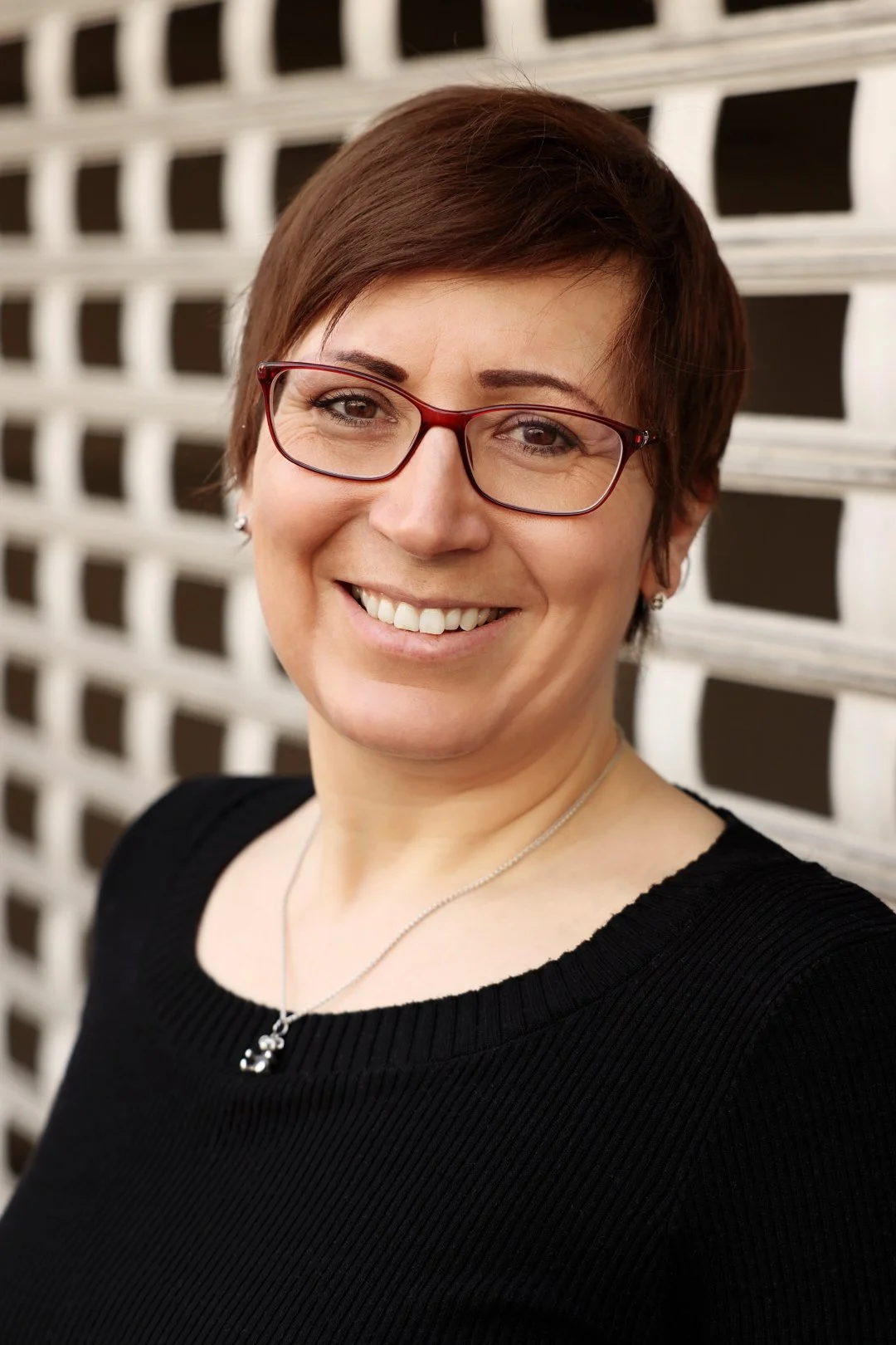 Nahaufnahme einer lächelnden Frau mit kurz braunen Haaren, rotem Brillengestell und schwarzem Oberteil vor einem weißen Gitter-Hintergrund.