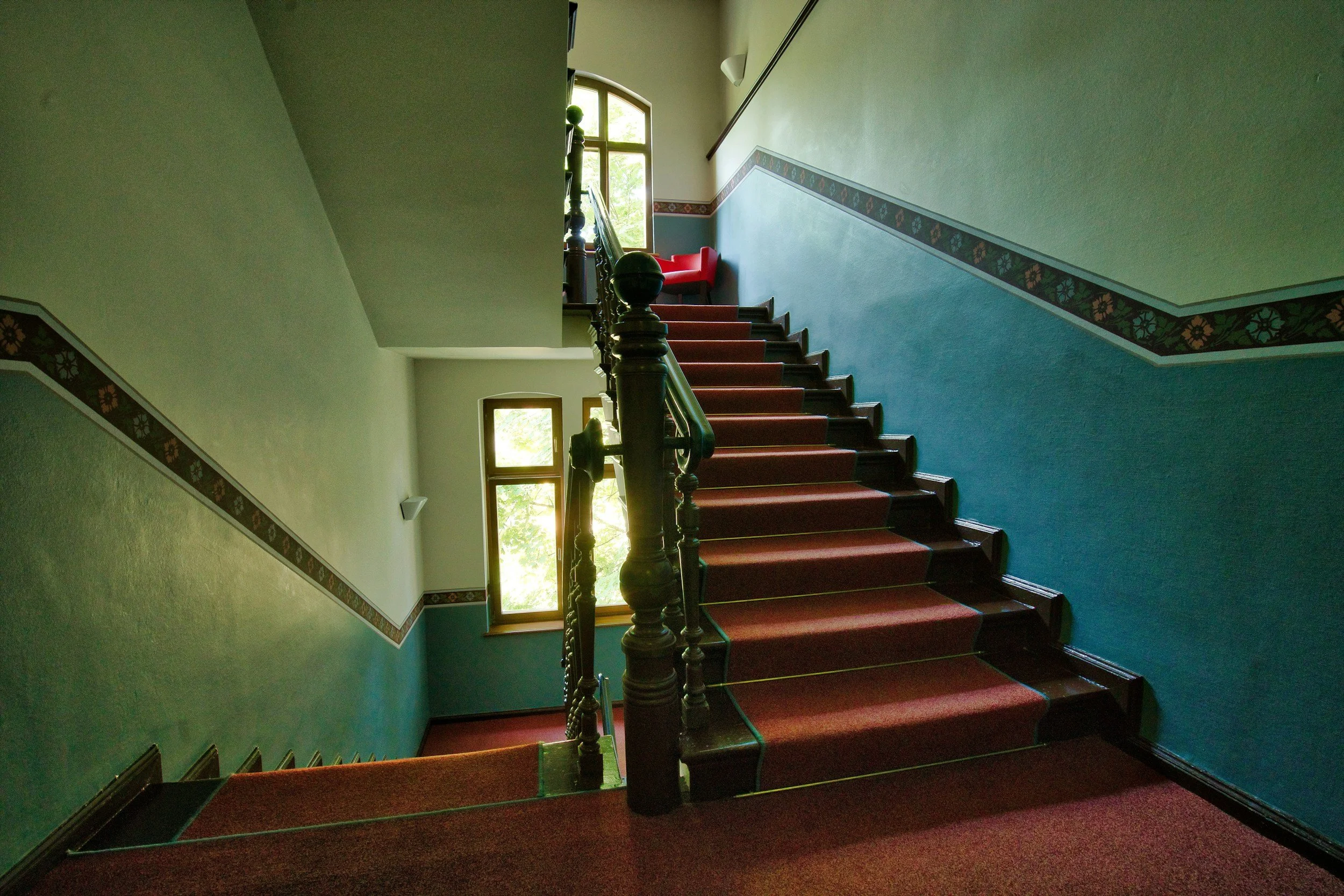 Indoor staircase with red carpet, dark wood railing, teal and cream walls, and windows letting in natural light.