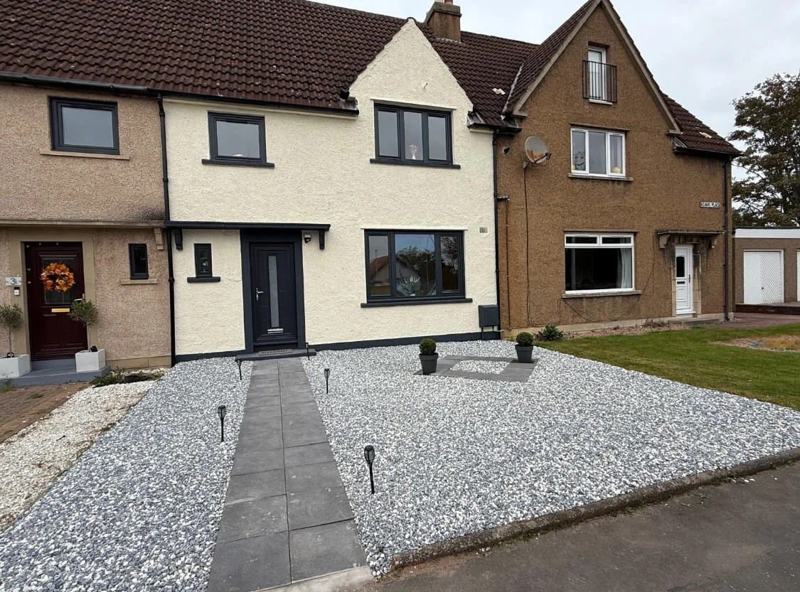 Front view of a row house with a pebble stone yard, gray walkway, and two potted plants. The house has a black front door and black-framed windows.