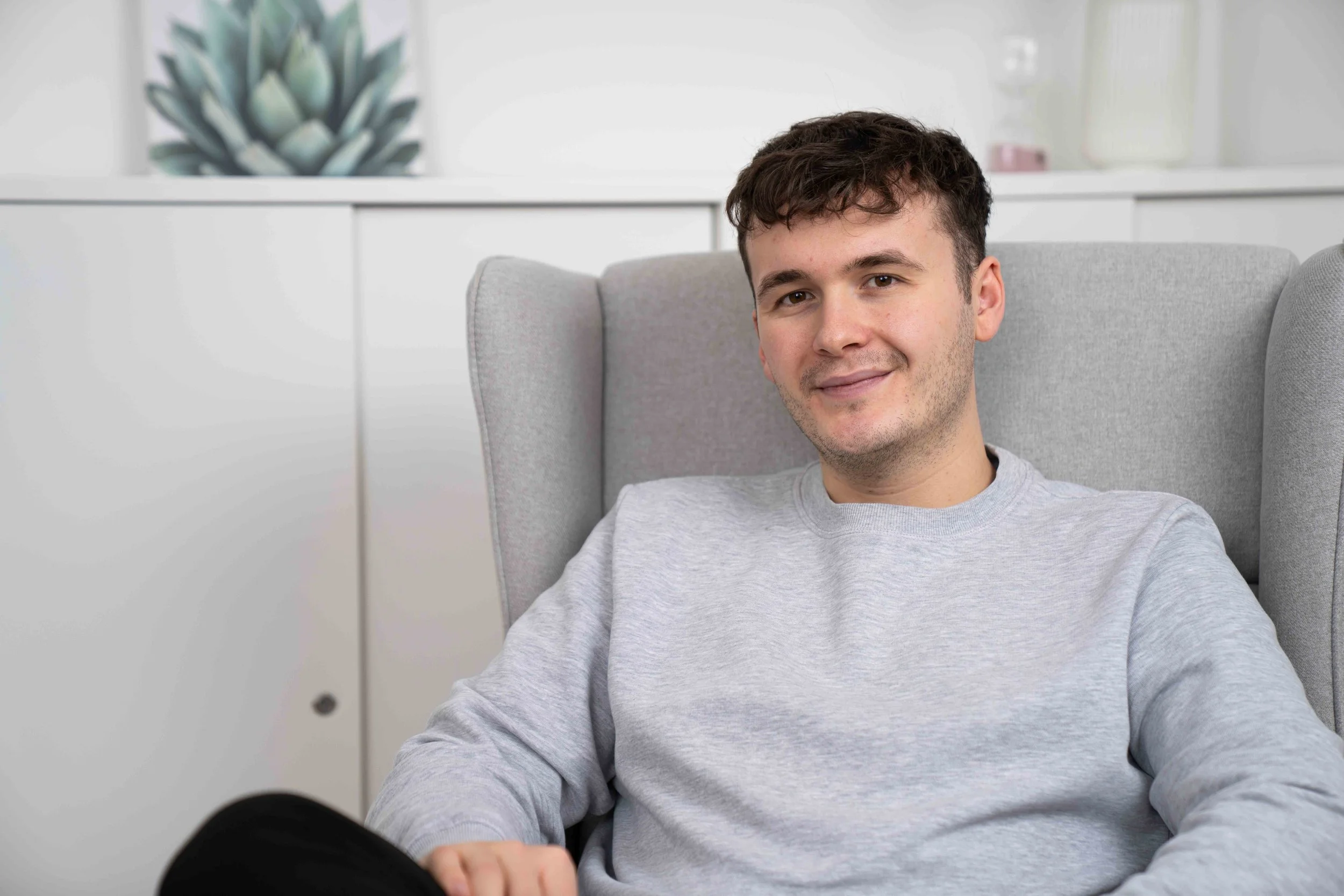 Der Therapeut sitzt entspannt in einem grauen Sessel in einem modernen, weißen Raum und lächelt.