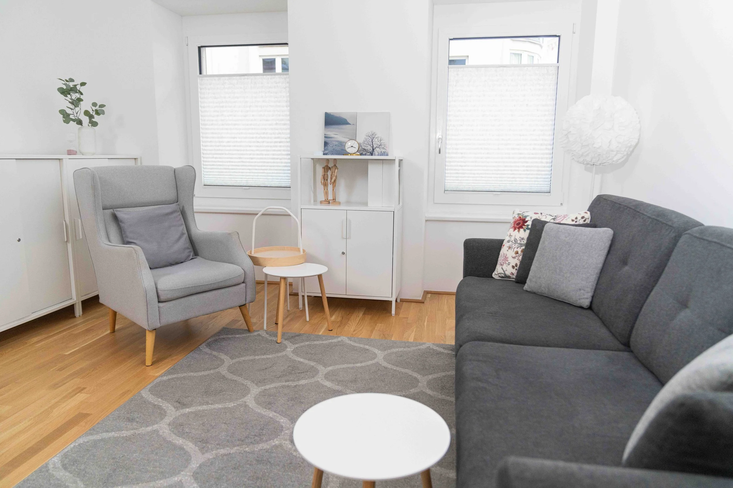 Helles Wohnzimmer mit grauer Couch, grauem Armchair, weißen Wänden, zwei Fenstern mit Rollos, weißen Möbeln, Holzfußboden, Teppich, Lampenschirm, Bilder und Dekorationsgegenständen.