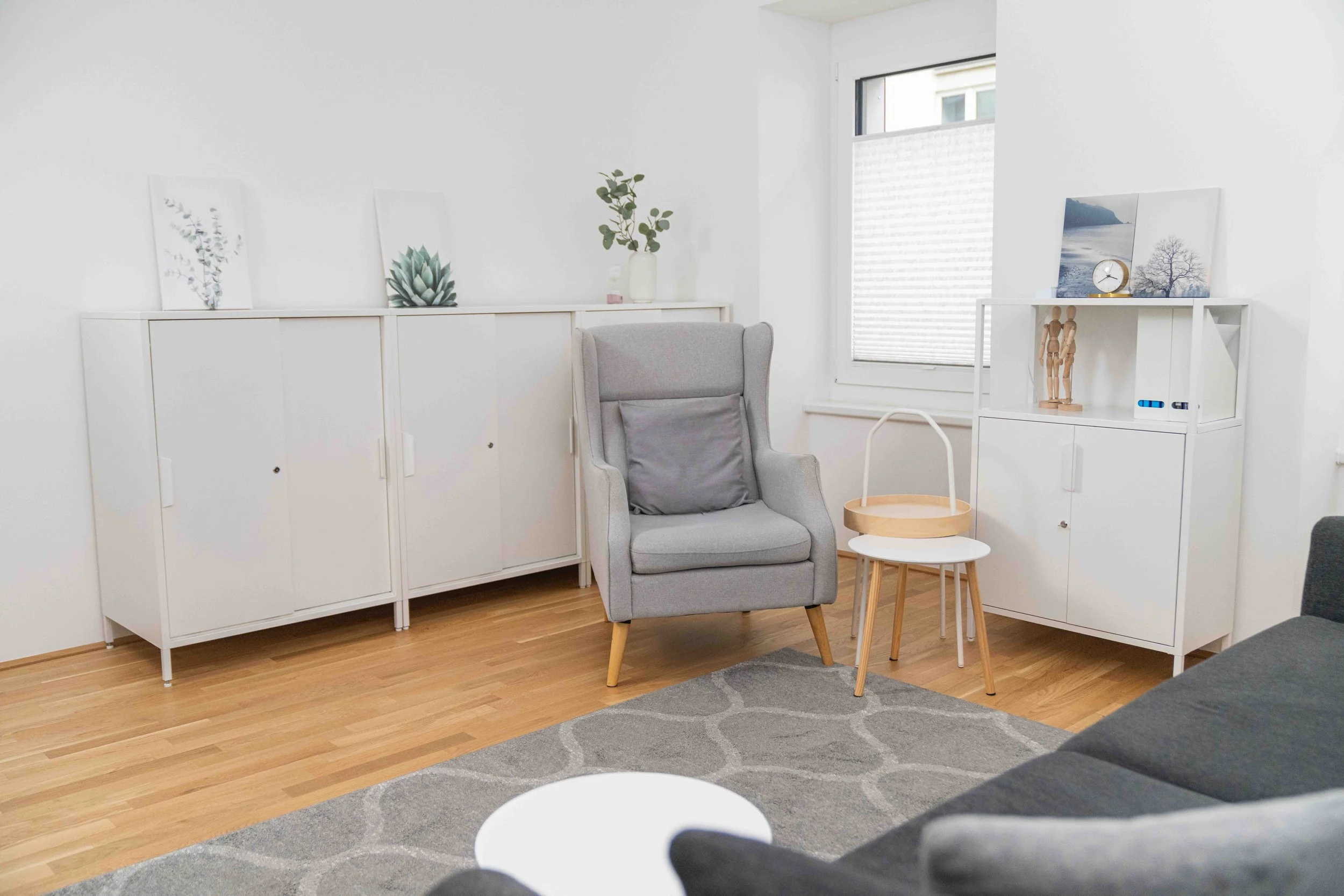 Ein modernes Wohnzimmer mit weißen Möbeln, einem grauen Sessel mit Kissen, einem kleinen Beistelltisch mit einem Topf, einem weißen Teppich und einer großen Fensterfront.