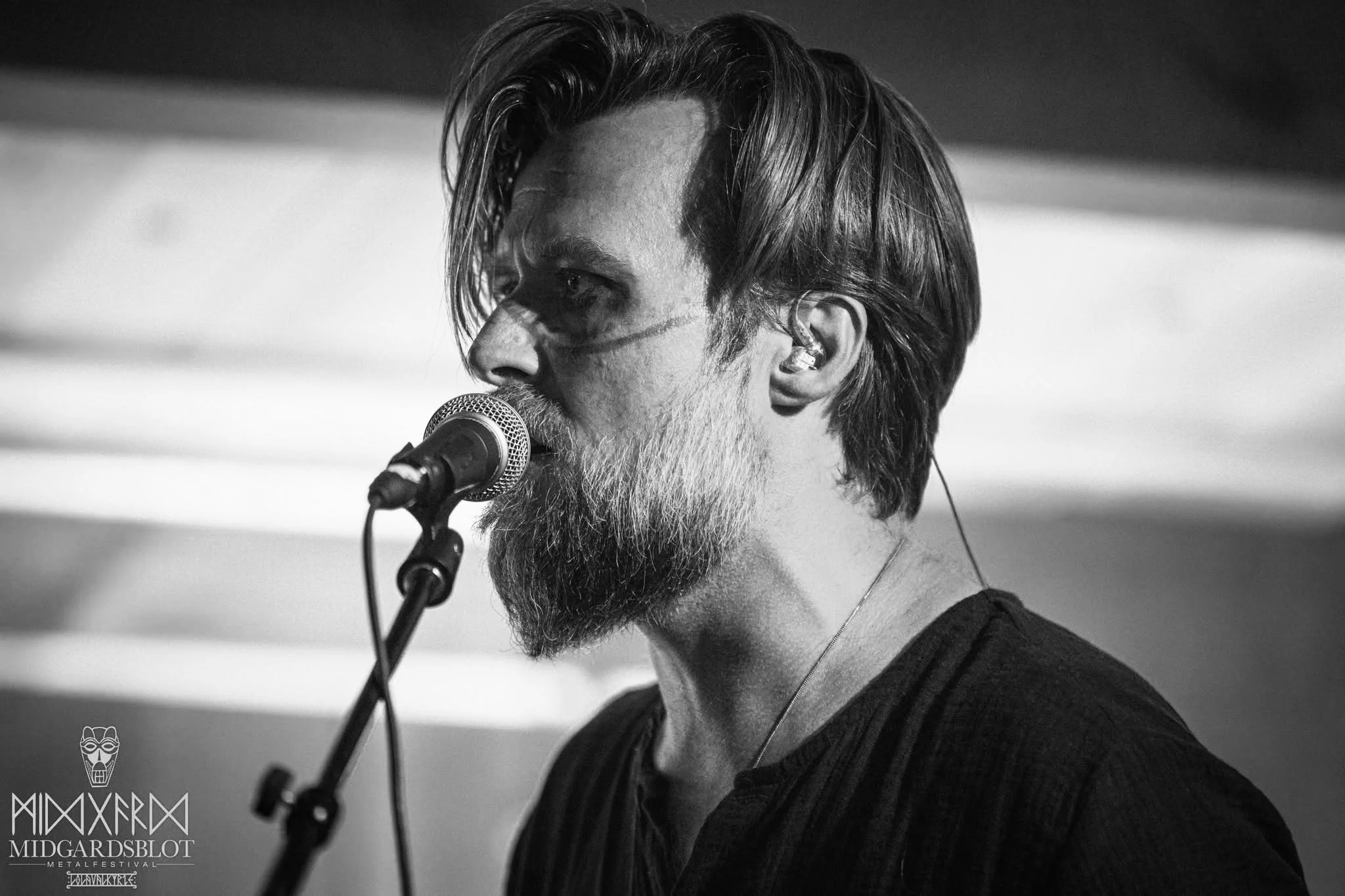 A black-and-white photo of a male musician singing or speaking into a microphone, with long hair, a beard, and wearing in-ear monitors. NEBALA concert at Midgårdsblot 2025. Jonas Lorentzen.
