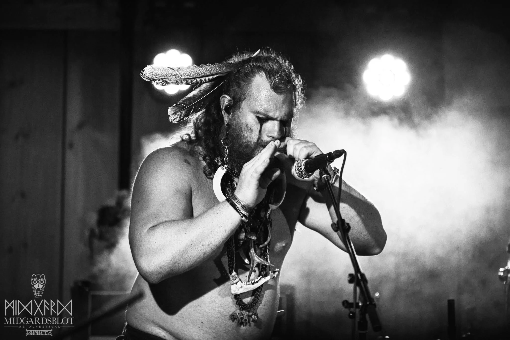 A shirtless man with long curly hair, wearing a feather headdress, playing a wind instrument on stage with bright backlights and smoke. NEBALA concert at Midgårdsblot 2025. Jonas Lorentzen. Ghili Prati.
