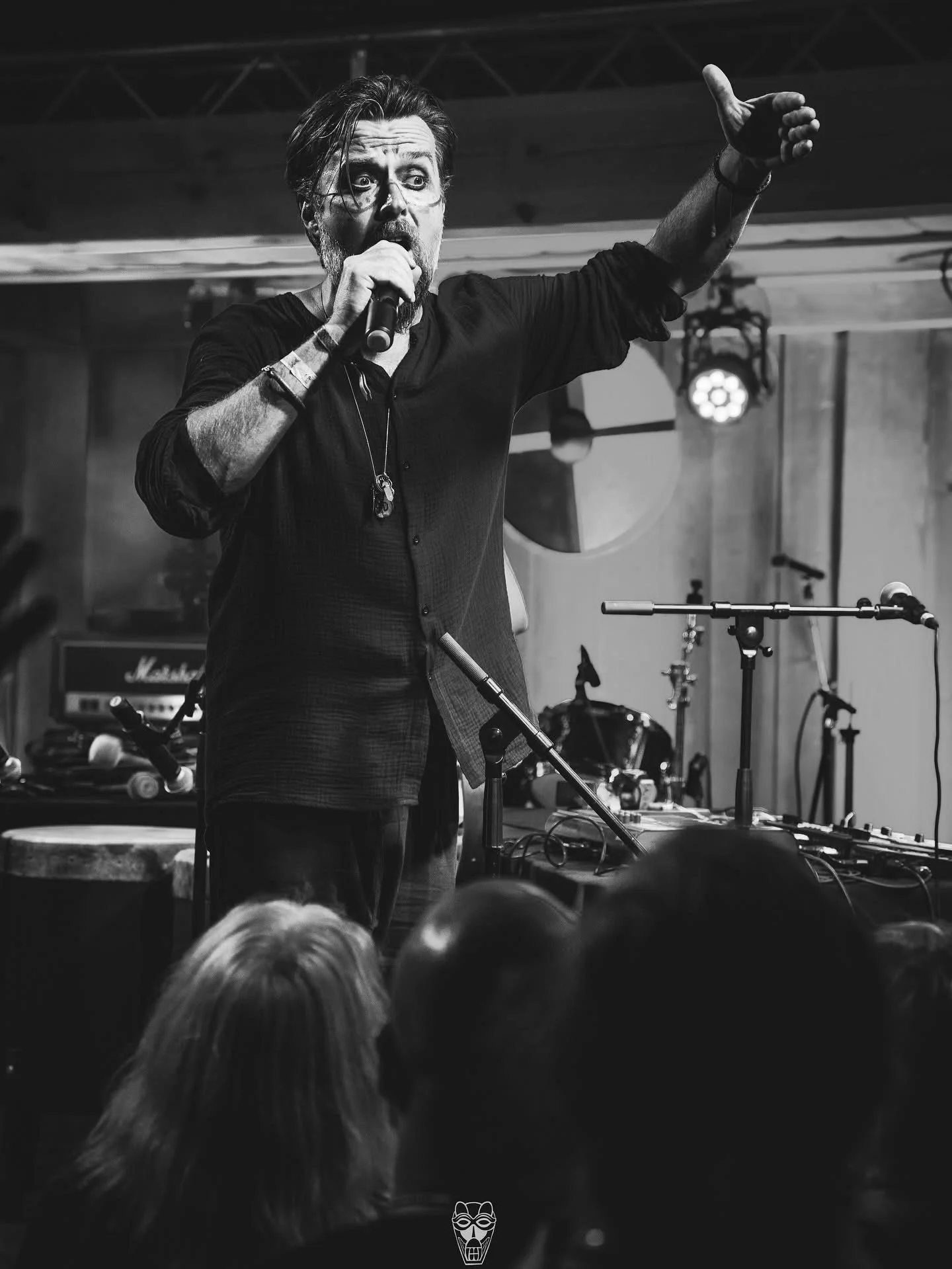 Black and white photo of a male singer performing on stage, holding a microphone with his left hand and gesturing with his right hand. Audience members' heads are visible in the foreground. Musical equipment can be seen in the background. NEBALA