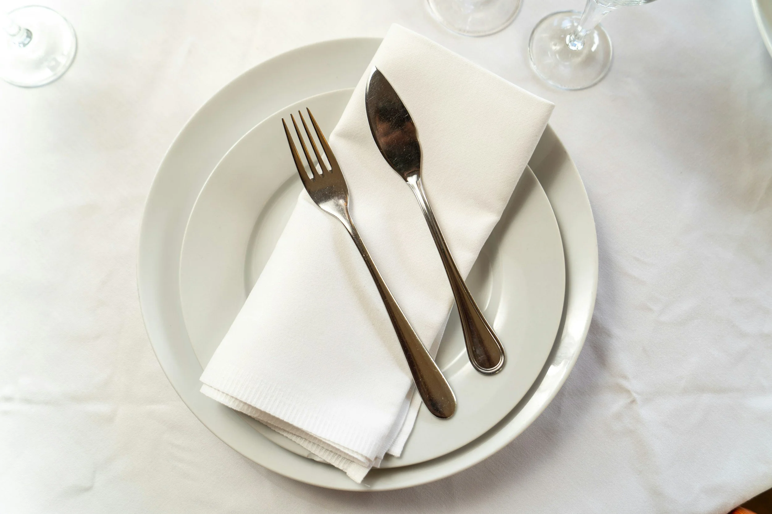 Place setting with a white plate, a white napkin, a fork, and a knife on a white tablecloth, with empty wine glasses in the background.