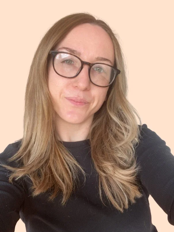 A woman with long, wavy blonde hair wearing glasses and a black top, smiling softly at the camera against a plain beige background.