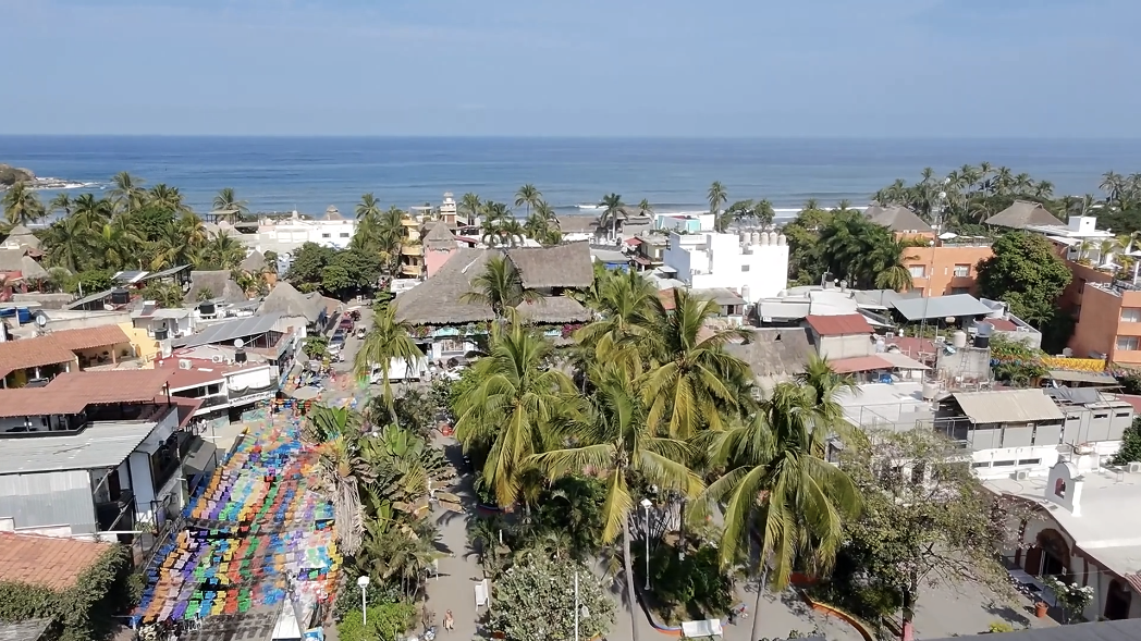 A coastal town with colorful market stalls, palm trees, and buildings, overlooking the ocean under a partly cloudy sky.