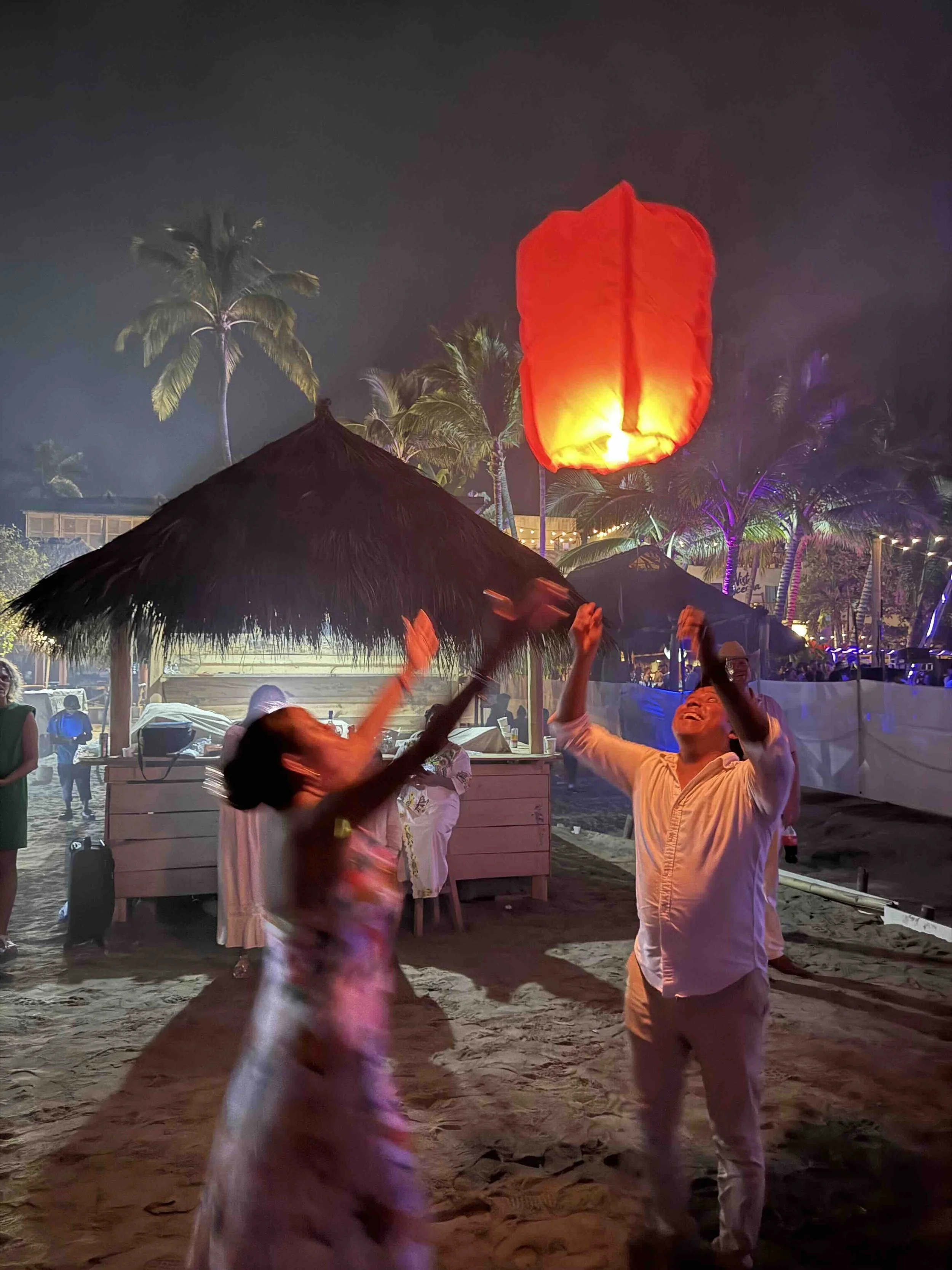 People releasing a red sky lantern on a beach at night, with palm trees and straw umbrellas in the background.