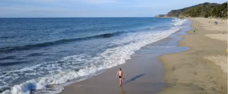 Person walking along a sandy beach by the ocean with waves, cliffs in the distance, and a few beach umbrellas.