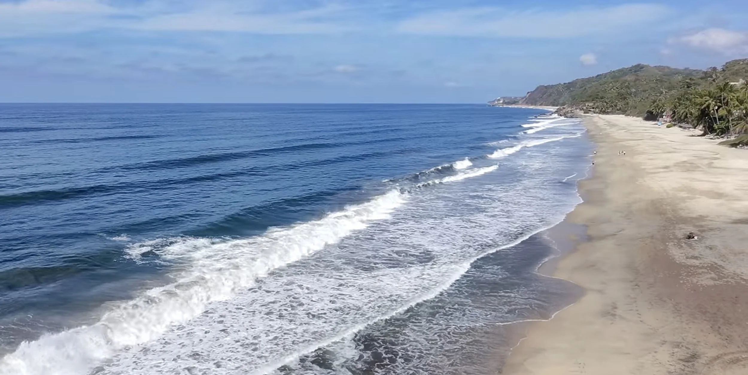 A view of a sandy beach with blue ocean waves under a partly cloudy sky, with green hills in the distance.