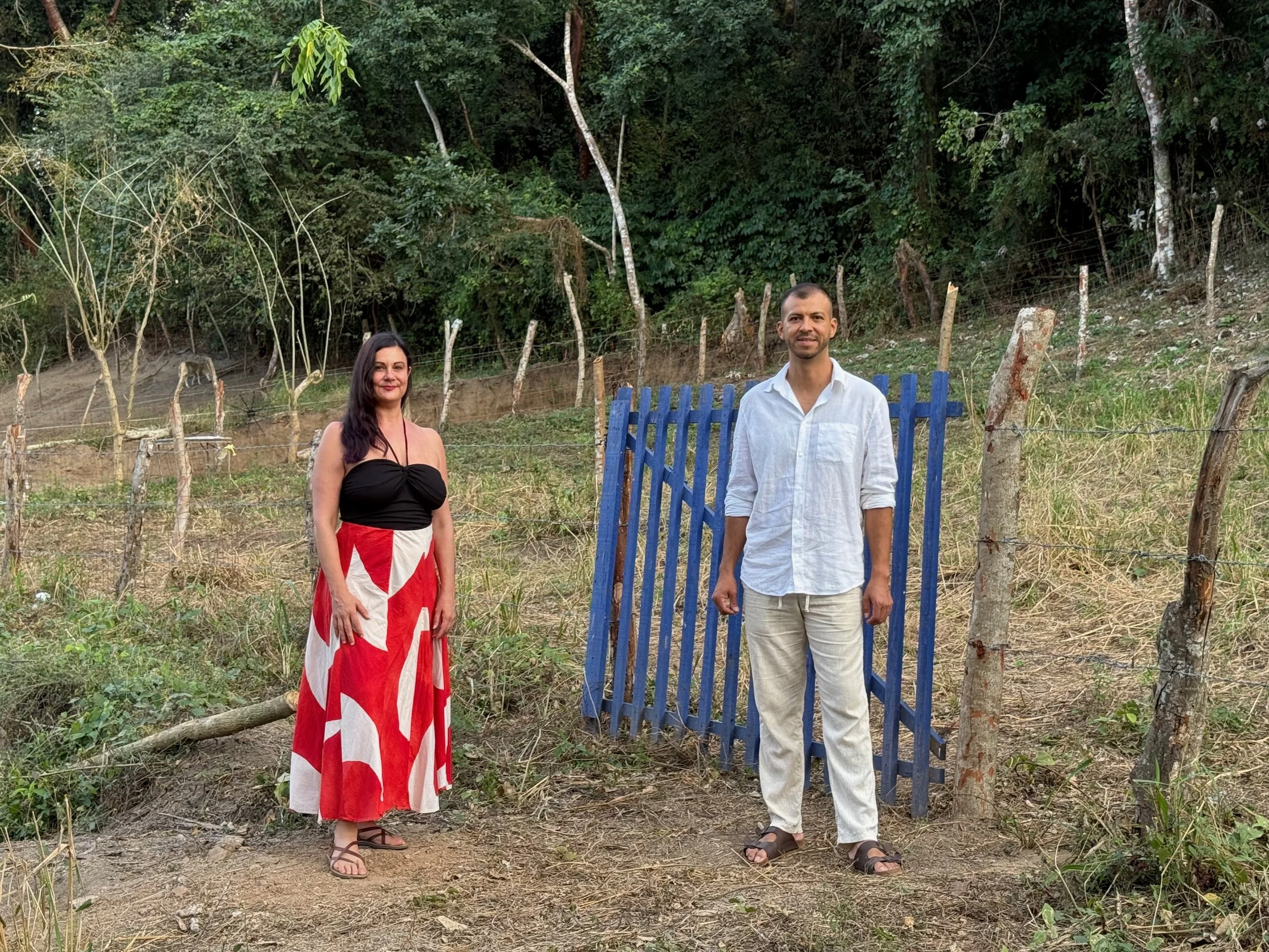A woman and a man standing outdoors near a blue gate in a rural area with a wooded hillside in the background. The woman wears a black top and a red and white patterned skirt, the man wears a white shirt and light-colored pants.