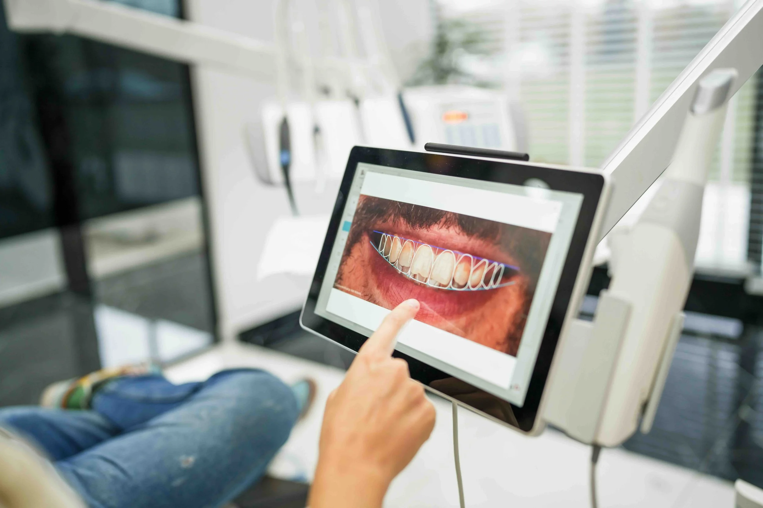 A person sitting on a chair at a dental clinic, pointing at an image of teeth on a tablet, with dental tools on a tray in the background.