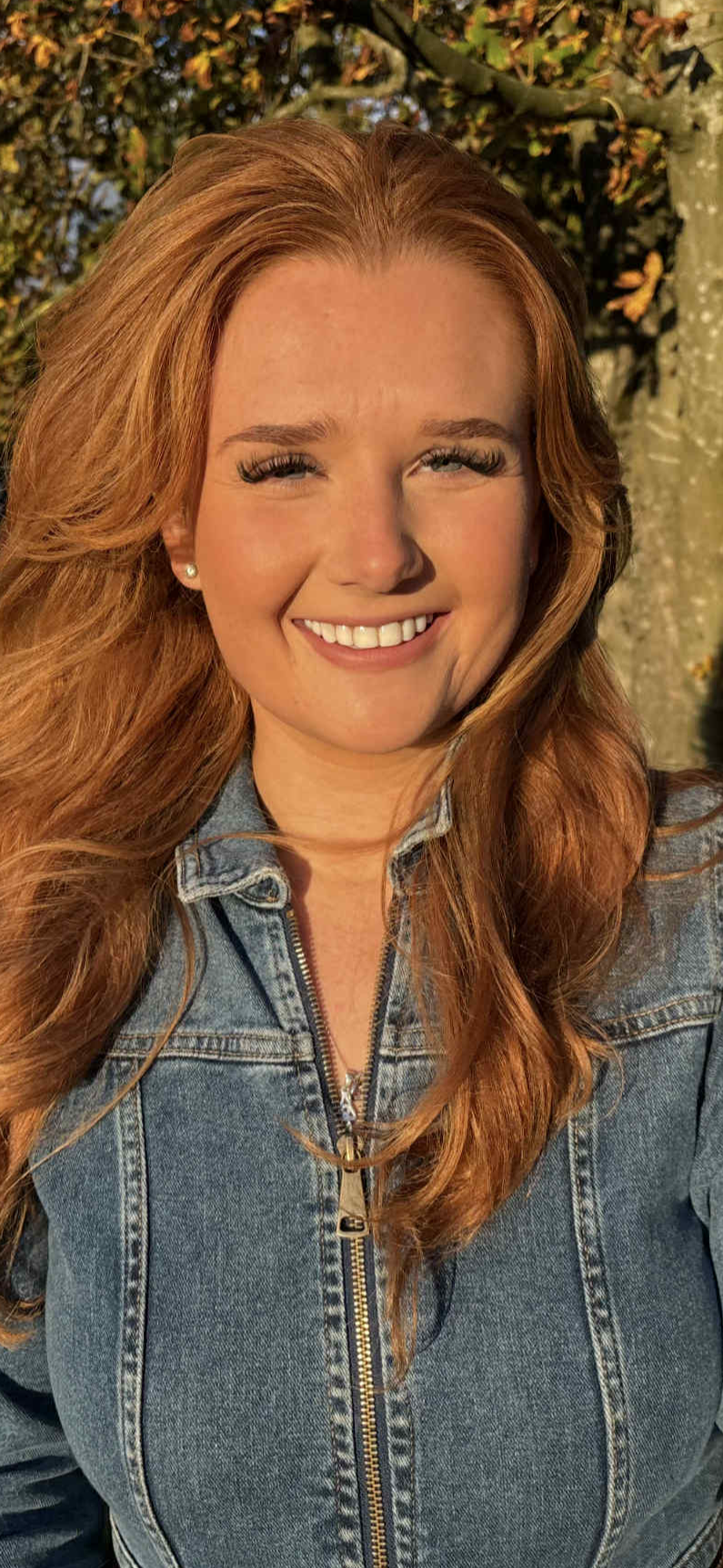A woman with long red hair smiling outdoors wearing a denim jacket, with trees and sunlight in the background.