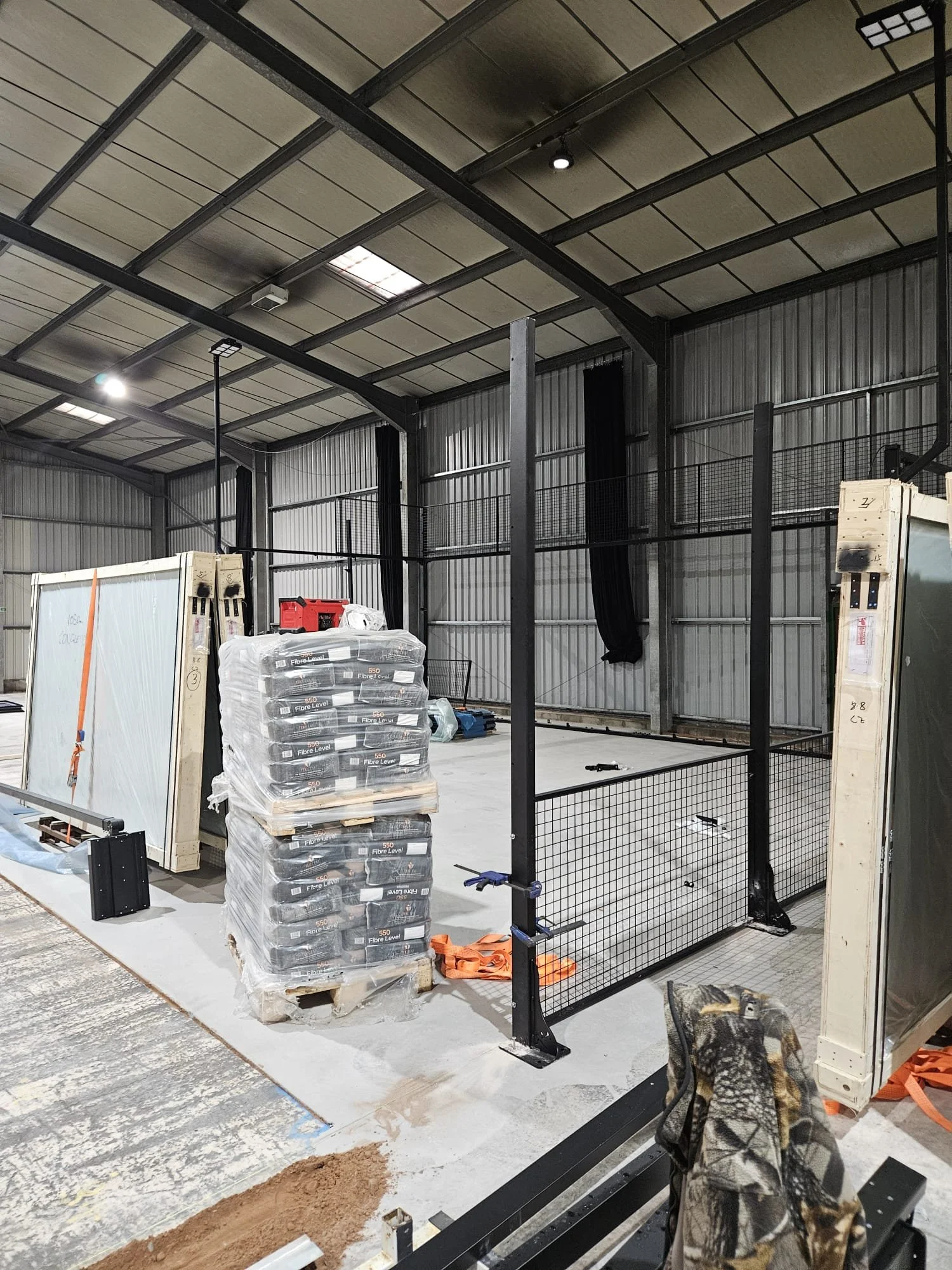 Construction site inside a warehouse with metal walls and roof, showing ongoing build of a structure with black metal framing, stacked wrapped pallets, and construction materials on the ground.