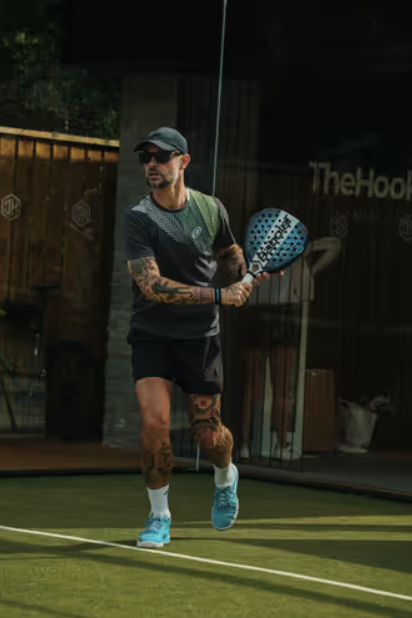A man with tattoos wearing sunglasses, a cap, black shorts, and sneakers playing pickleball on an outdoor court.