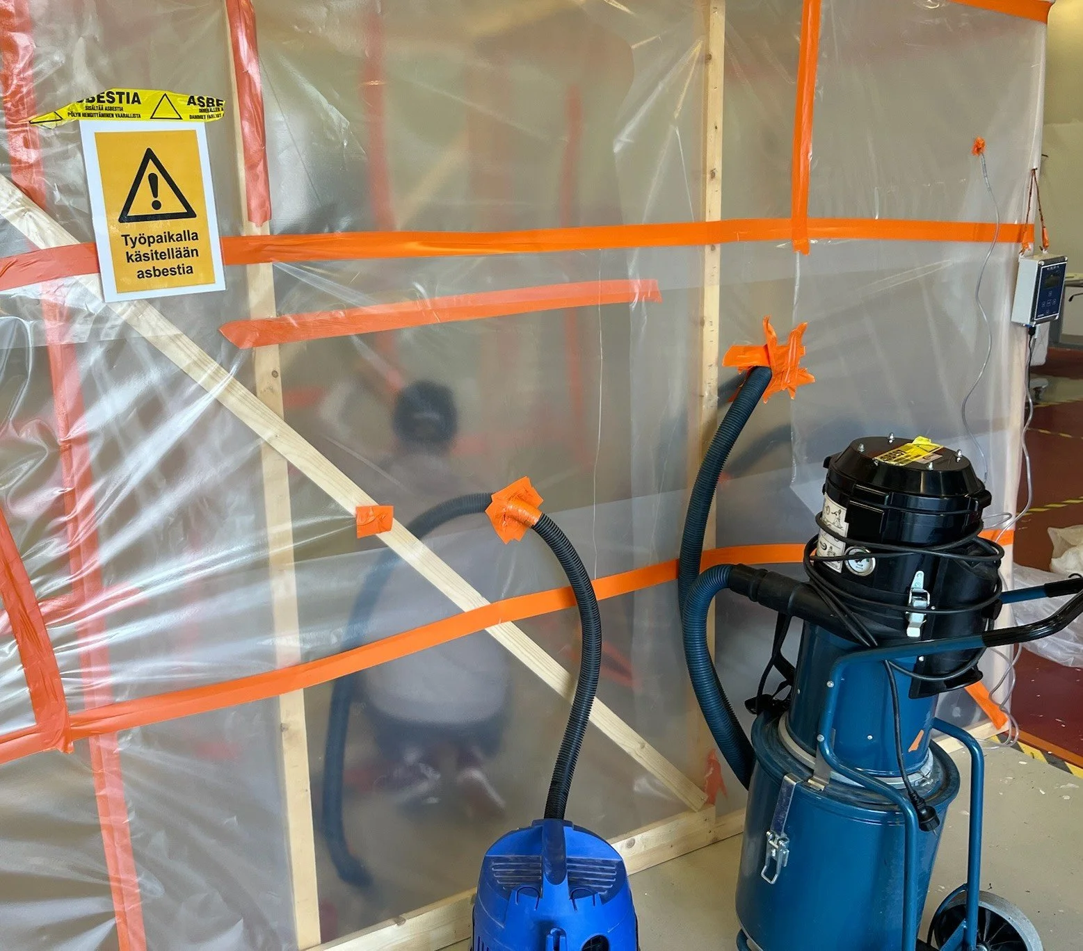 Varoitusmerkki, jossa lukee 'Työpaikalla käsitellään asbestia' ja rakennustarvikkeita, ilmastointilaitteita ja suojarakennelman osia rakennustyömaalla.