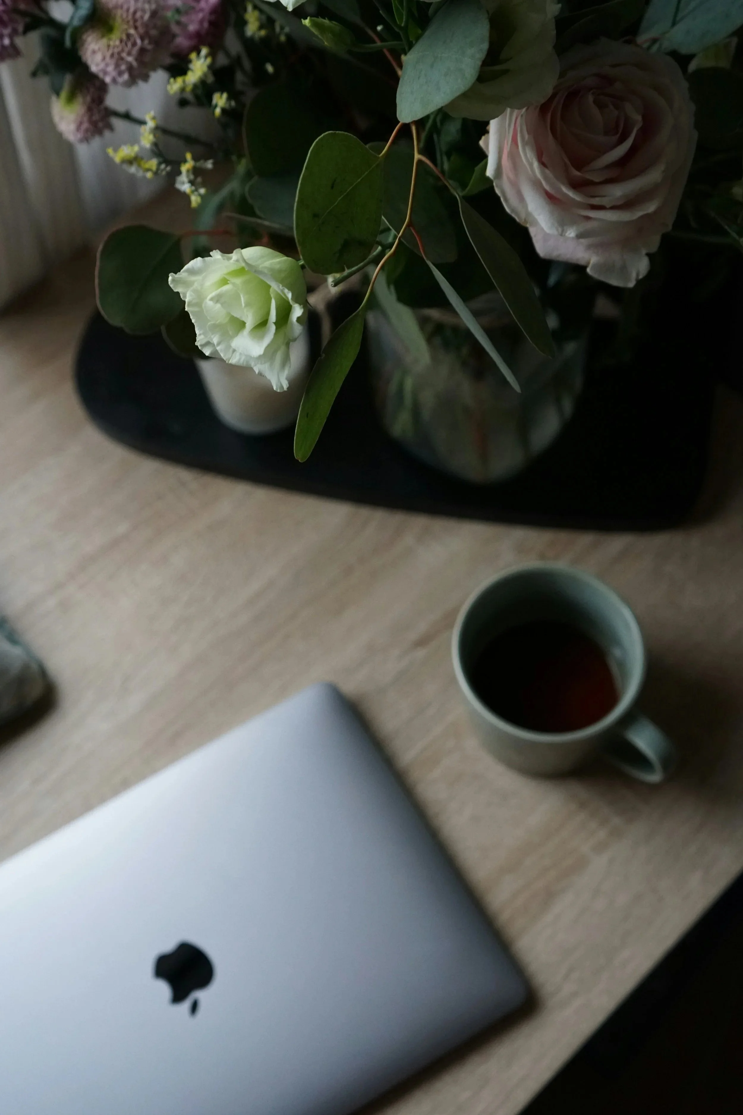 A wooden desk with a closed MacBook, a ceramic coffee mug with dark liquid, and a floral arrangement in a vase with green leaves and white and pink flowers.