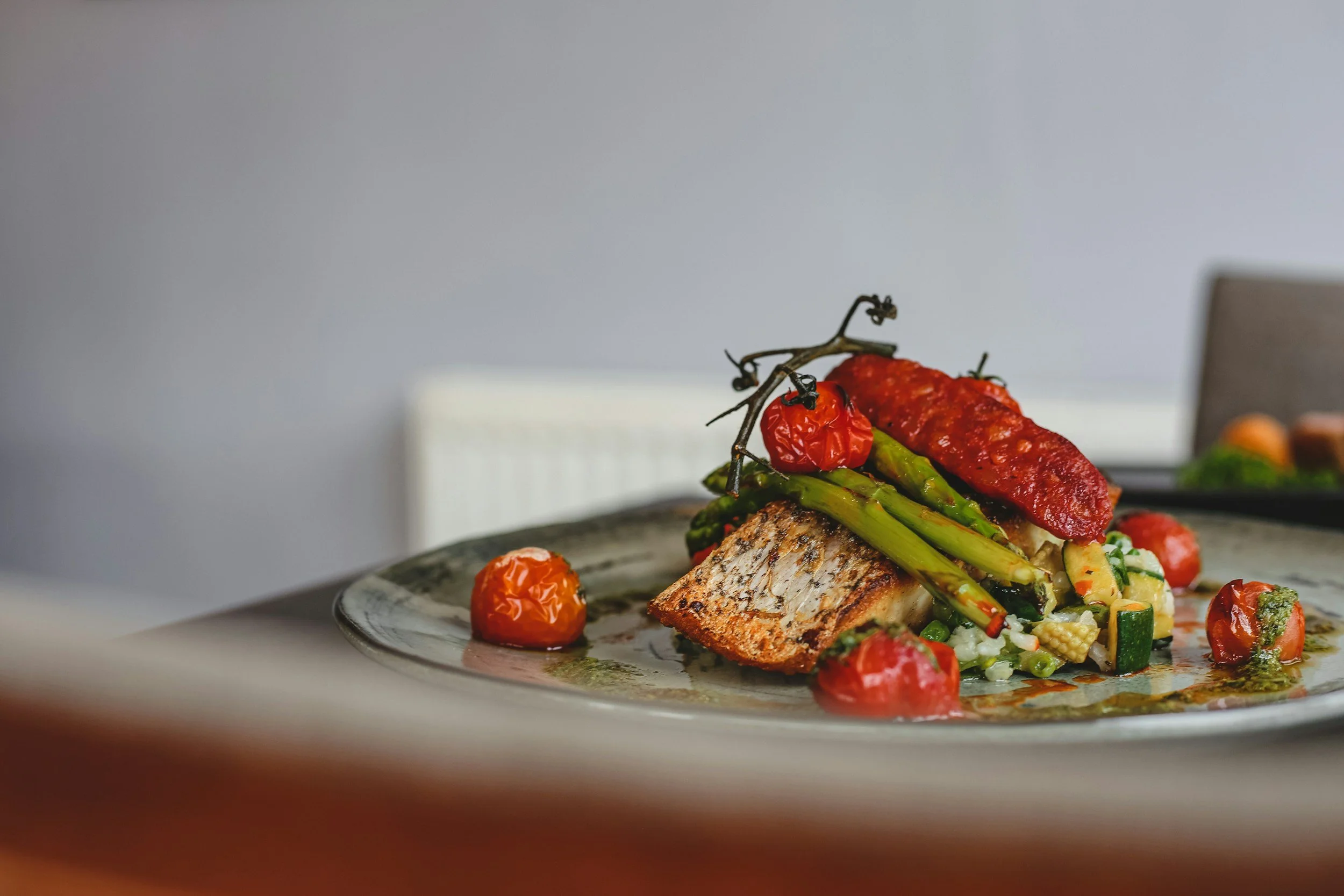 A plated dish featuring grilled fish with cherry tomatoes, asparagus, zucchini, baby corn, and a red sauce on top.