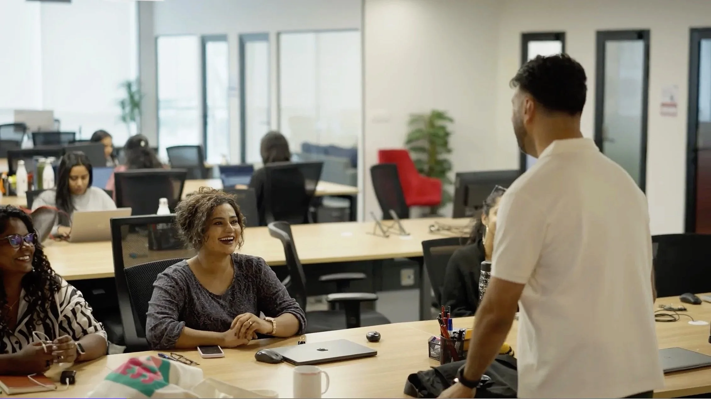 A group of diverse coworkers in a modern office conference room, with one standing and presenting to seated colleagues, who appear engaged and smiling.
