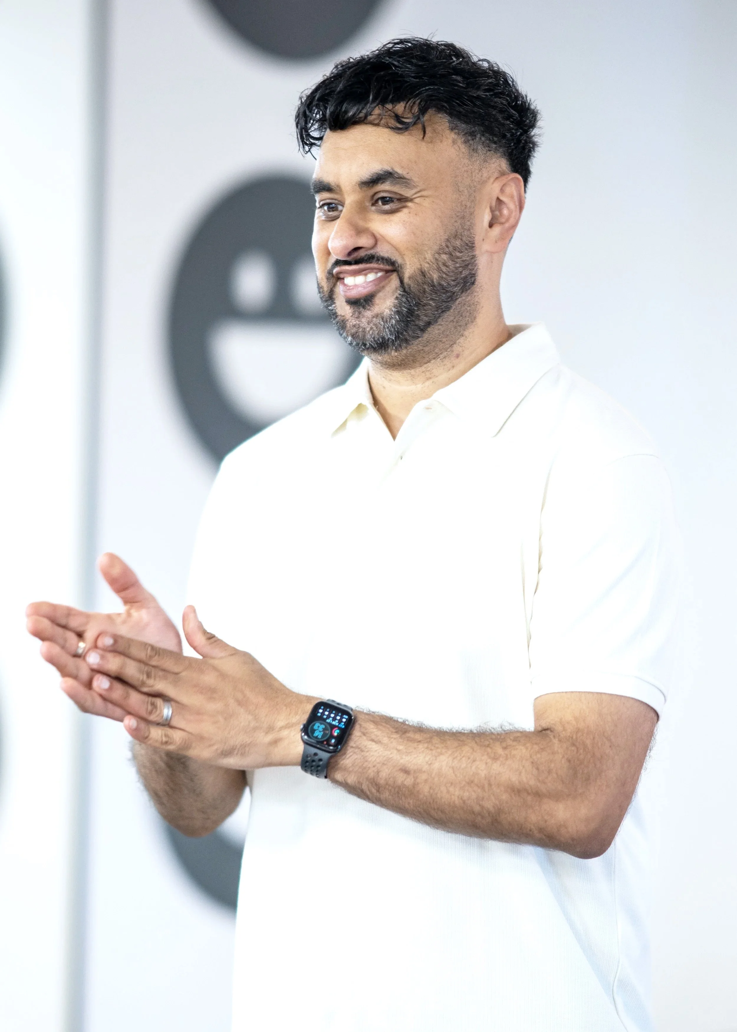 A man with dark hair, beard, and mustache, wearing a white polo shirt, claps his hands and smiles, standing in front of a blurred background with a black emoticon face.