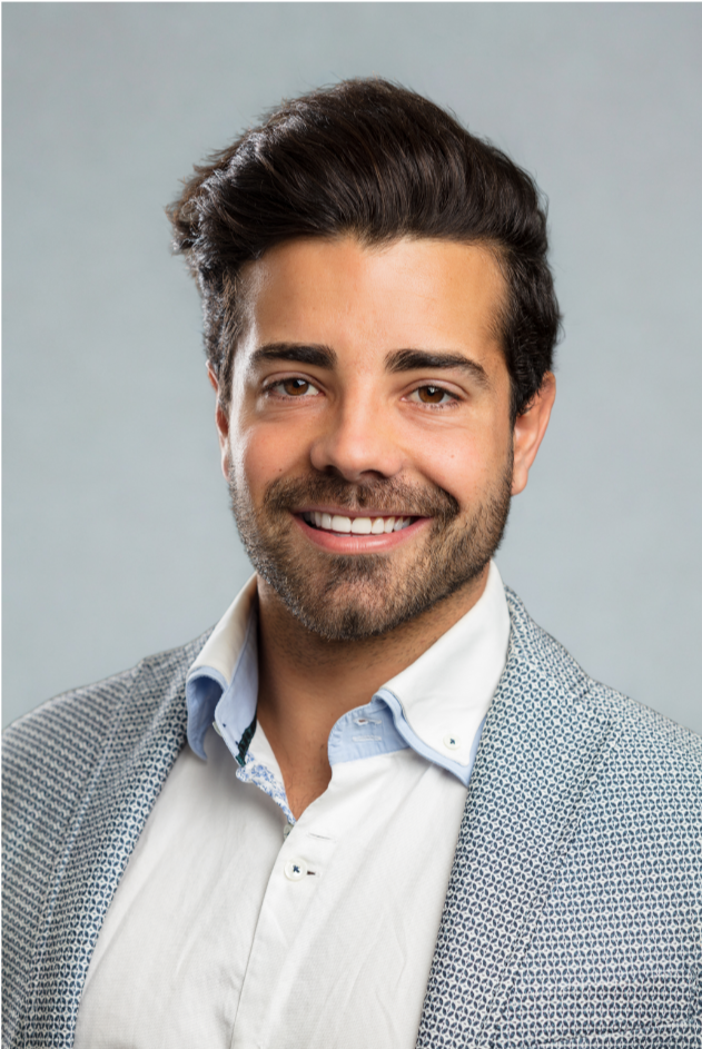 A man with dark hair, a beard, and a mustache, smiling in a professional headshot. He is wearing a white shirt and a patterned blazer, with a light gray background.