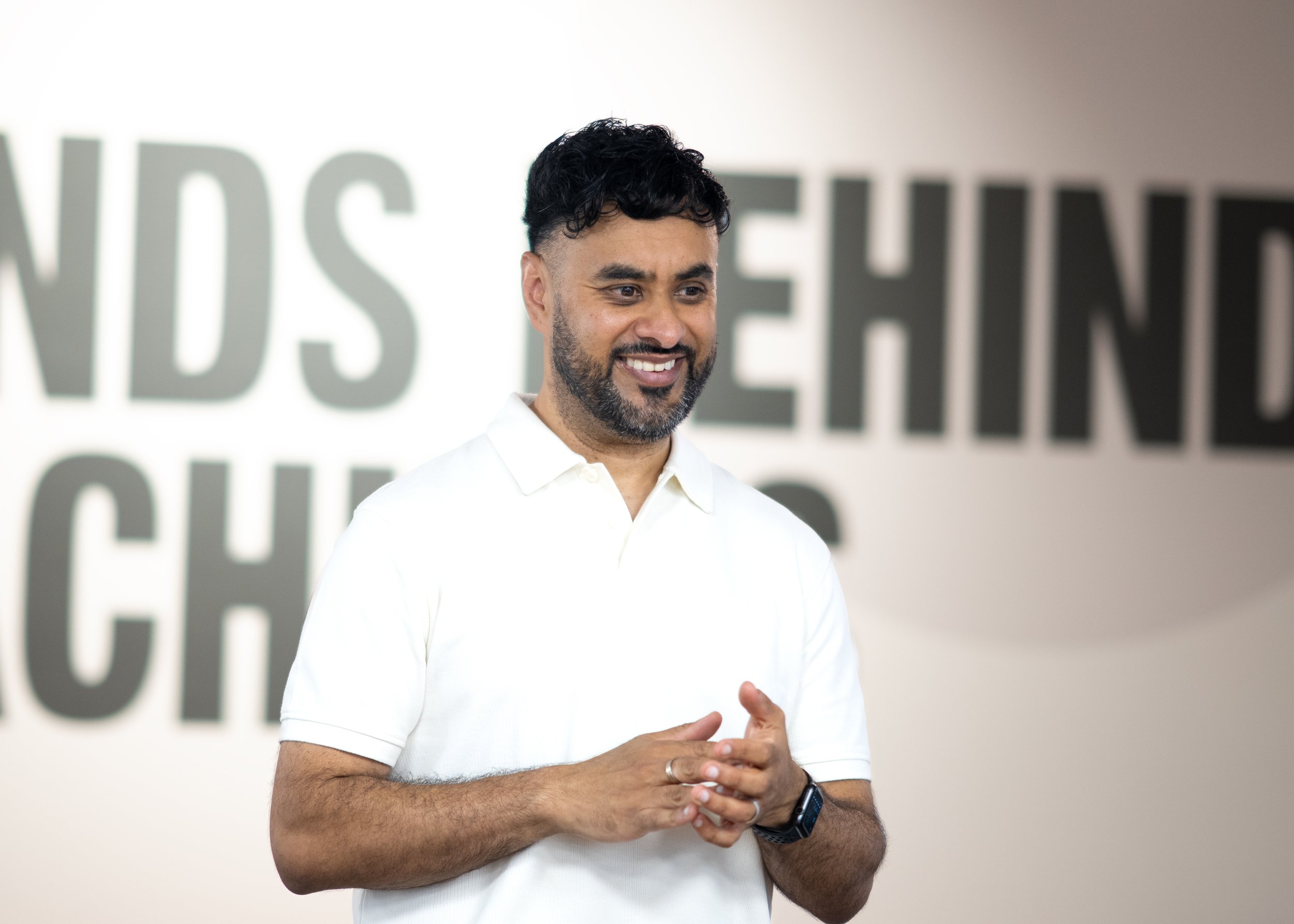 A man with dark hair and a beard smiling, wearing a white polo shirt and a smartwatch, standing in front of a wall with large text that is partially visible.