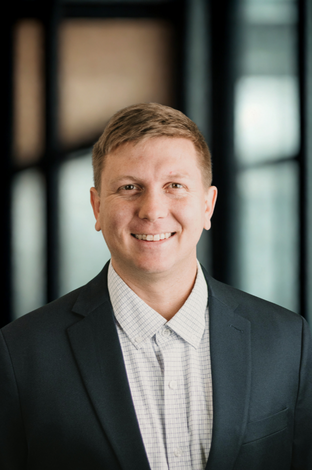 Professional portrait of a smiling man with short light brown hair, wearing a dark blazer and a light checkered shirt, in an office setting with large windows in the background.