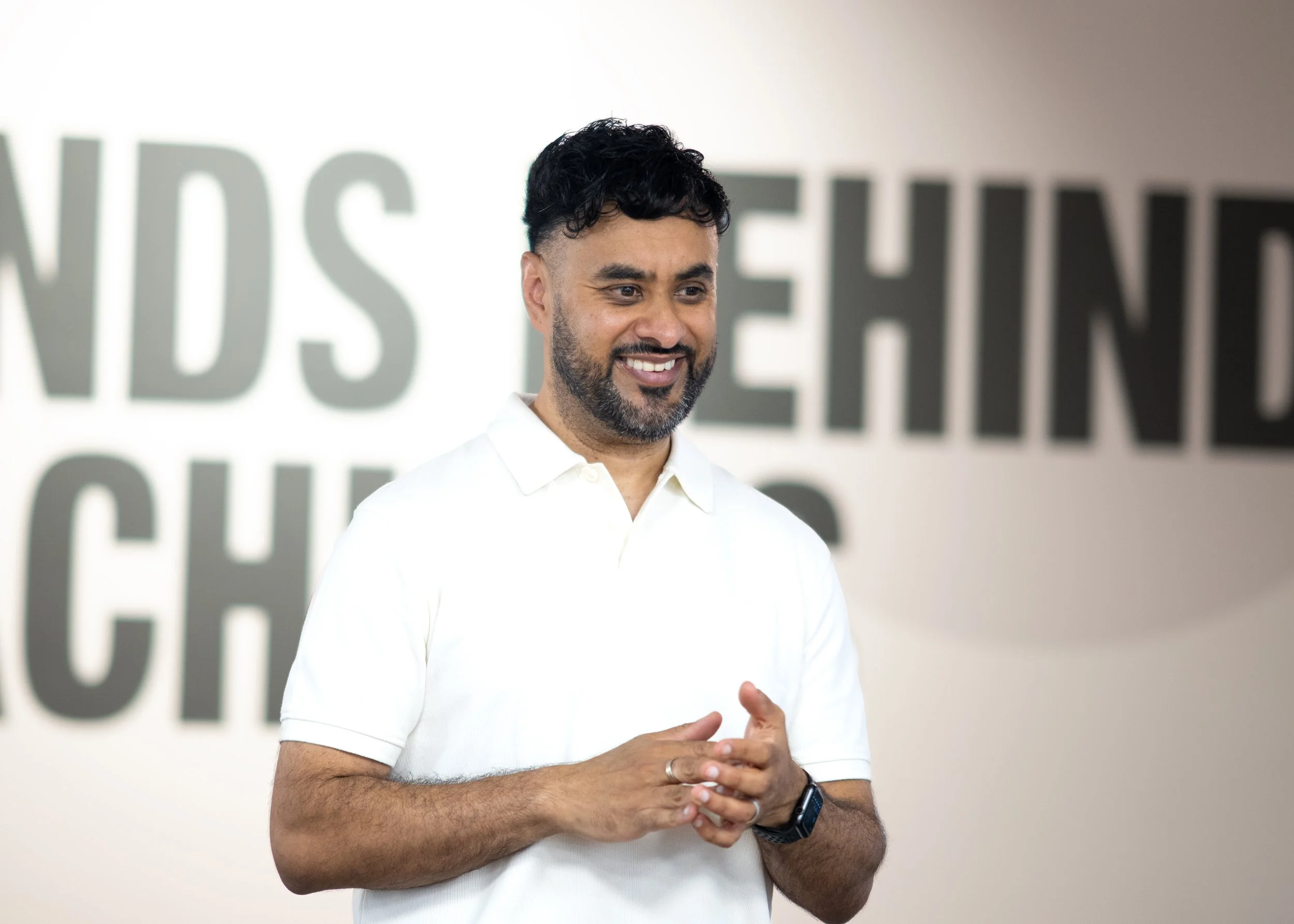 A man with dark hair and a beard wearing a white polo shirt is smiling and standing in front of a blurred background with large gray text.