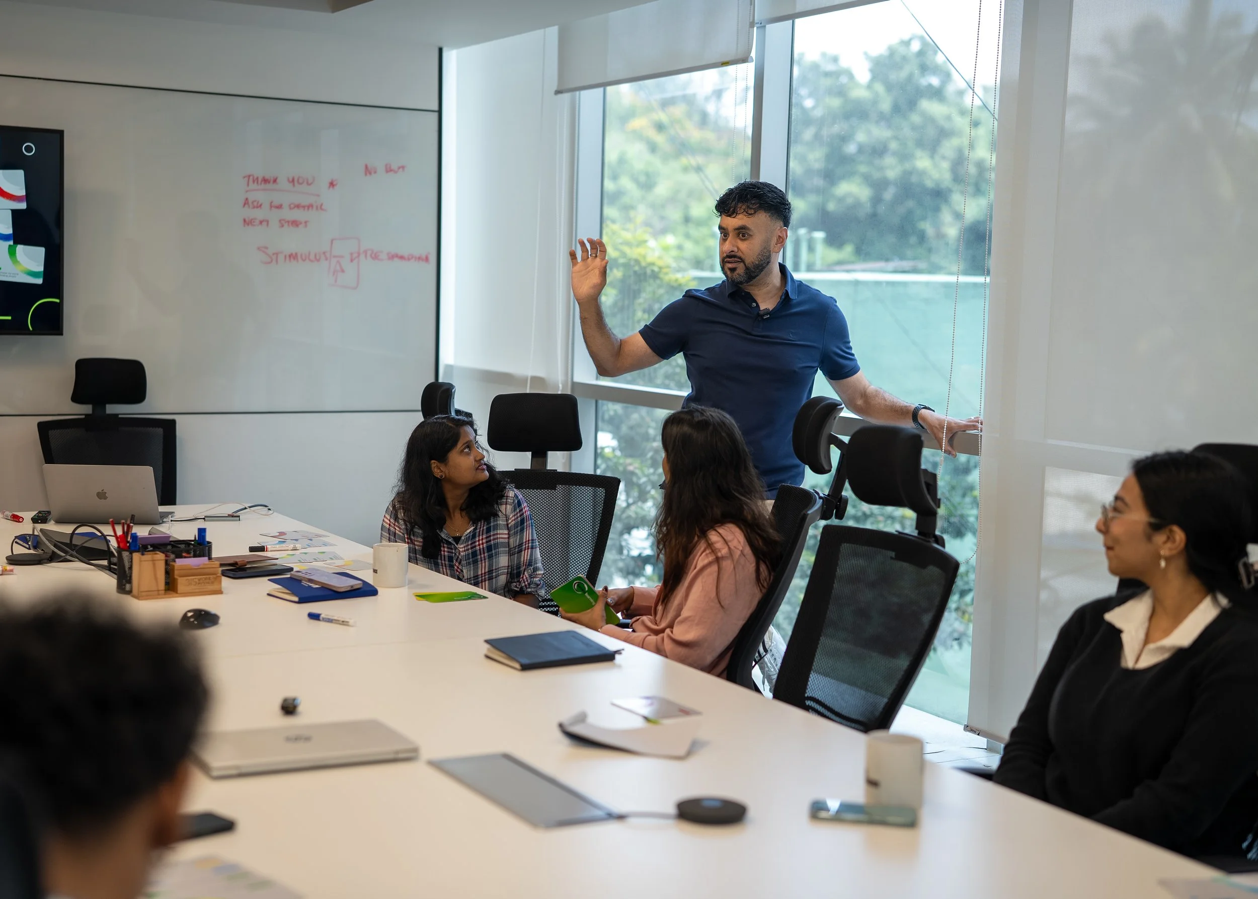 A man giving a presentation to a group of women in a modern conference room. The woman closest to the camera is looking up at him, while others are listening. There is a whiteboard with writing and a large window with natural light.