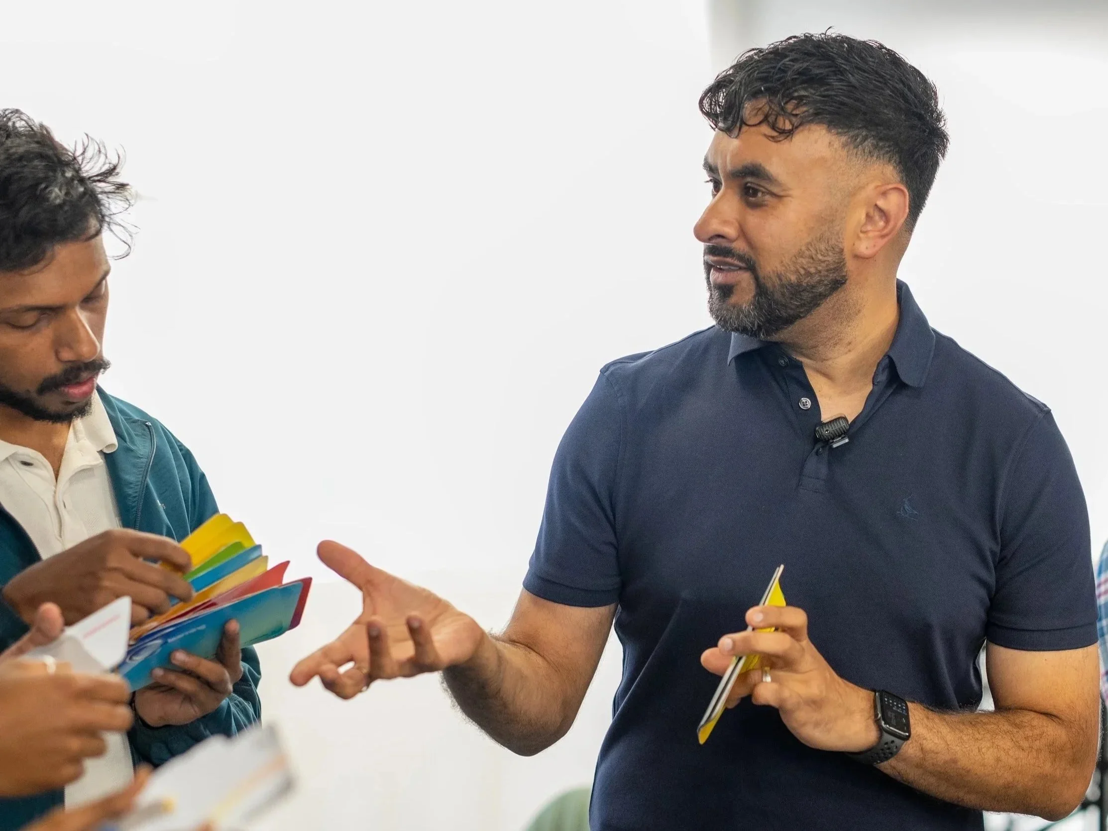 Two men engaged in conversation, with one man holding a yellow notepad and the other examining colorful cards.