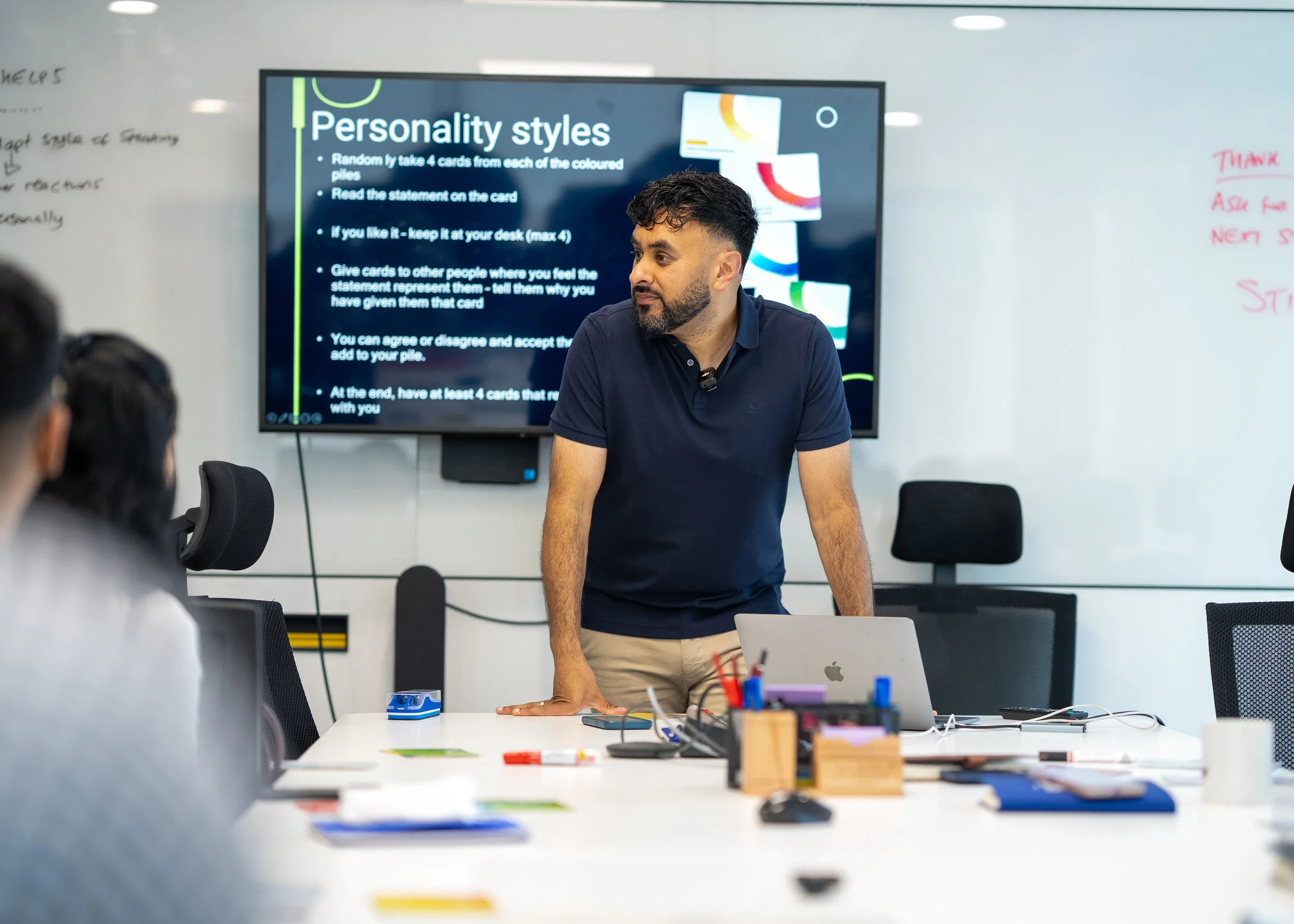 Man giving a presentation in a conference room with a large screen displaying a slide on personality styles, surrounded by colleagues and a whiteboard.
